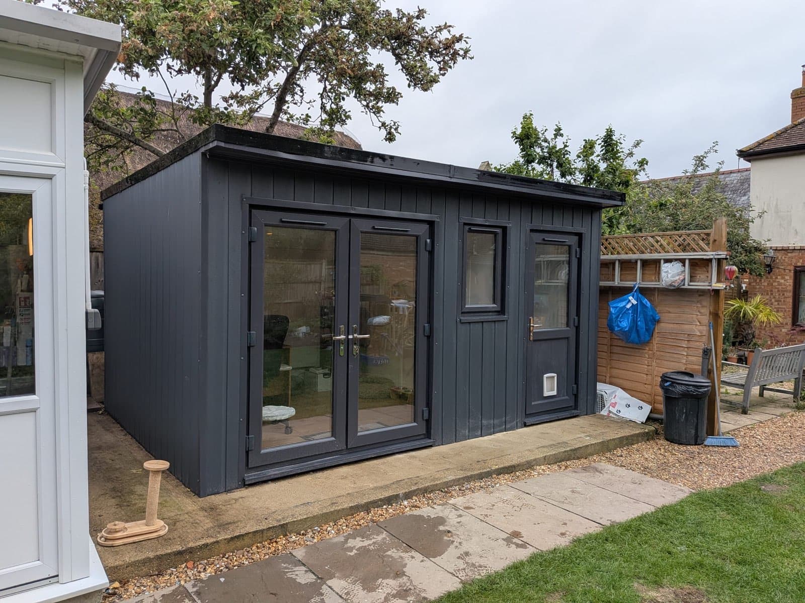 Anthracite grey composite-clad garden room with a flat roof, double glazed doors and two windows, standing on a paved base beside a lawn