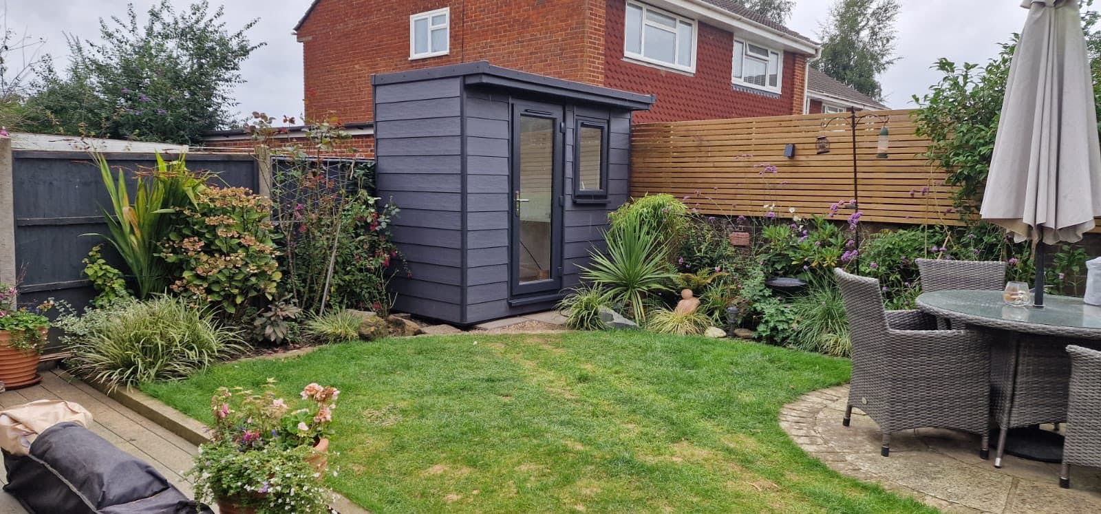 Compact anthracite garden office in the corner of a garden, with a patio, rattan dining set and parasol in front