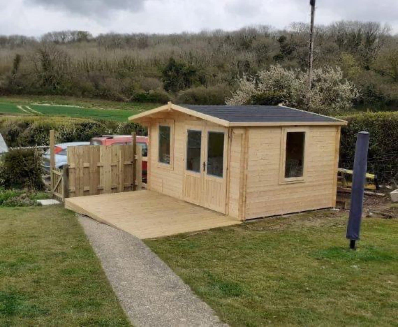 Untreated apex log cabin with a timber deck in front, in a rural field with hedgerows behind