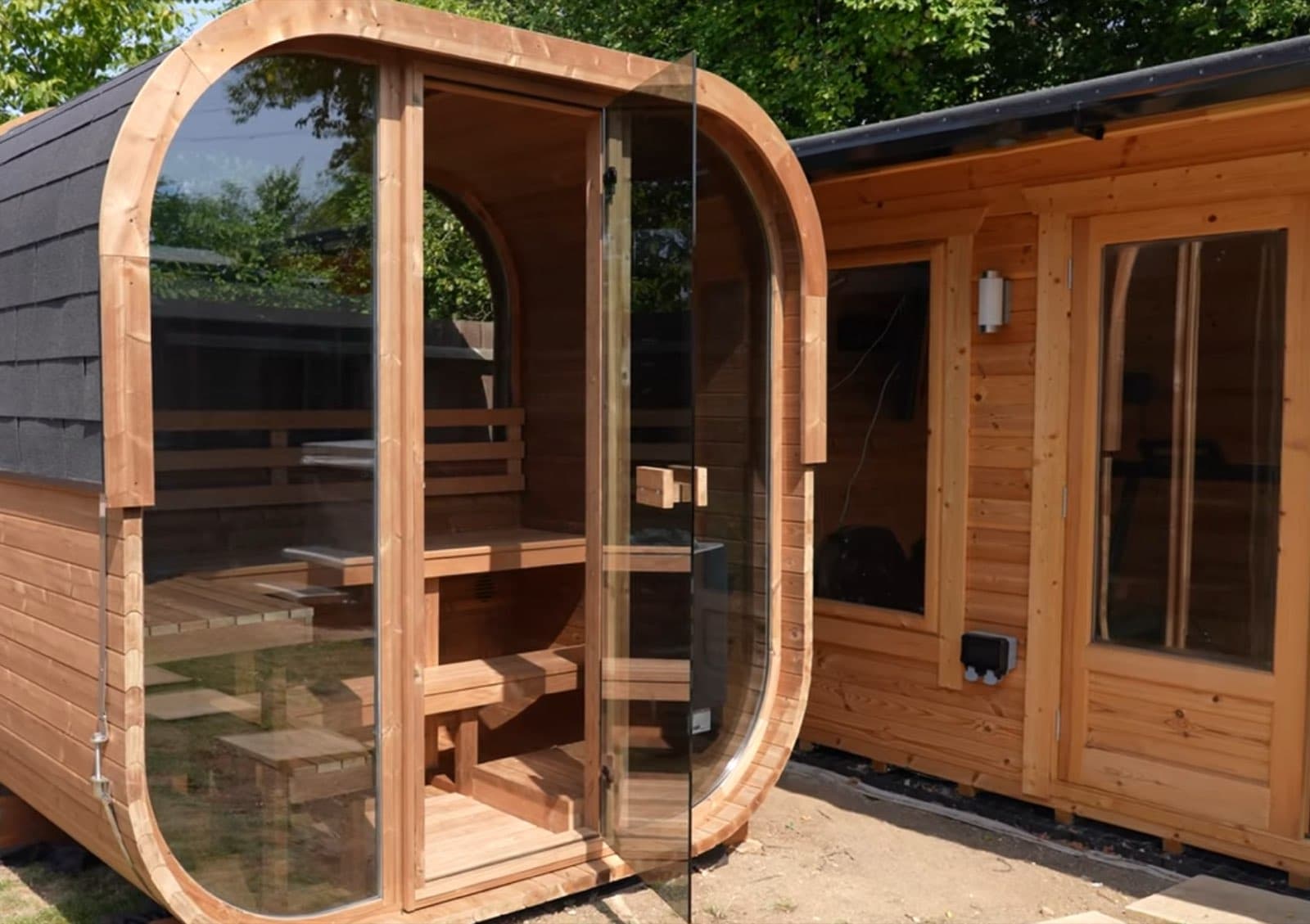 Barrel sauna with a curved glass door standing next to a log cabin, on display at the showroom