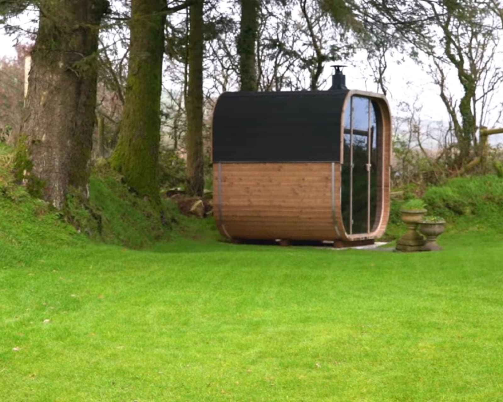 Barrel-shaped outdoor sauna with a charred black roof and a curved glass end, standing on a lawn in woodland