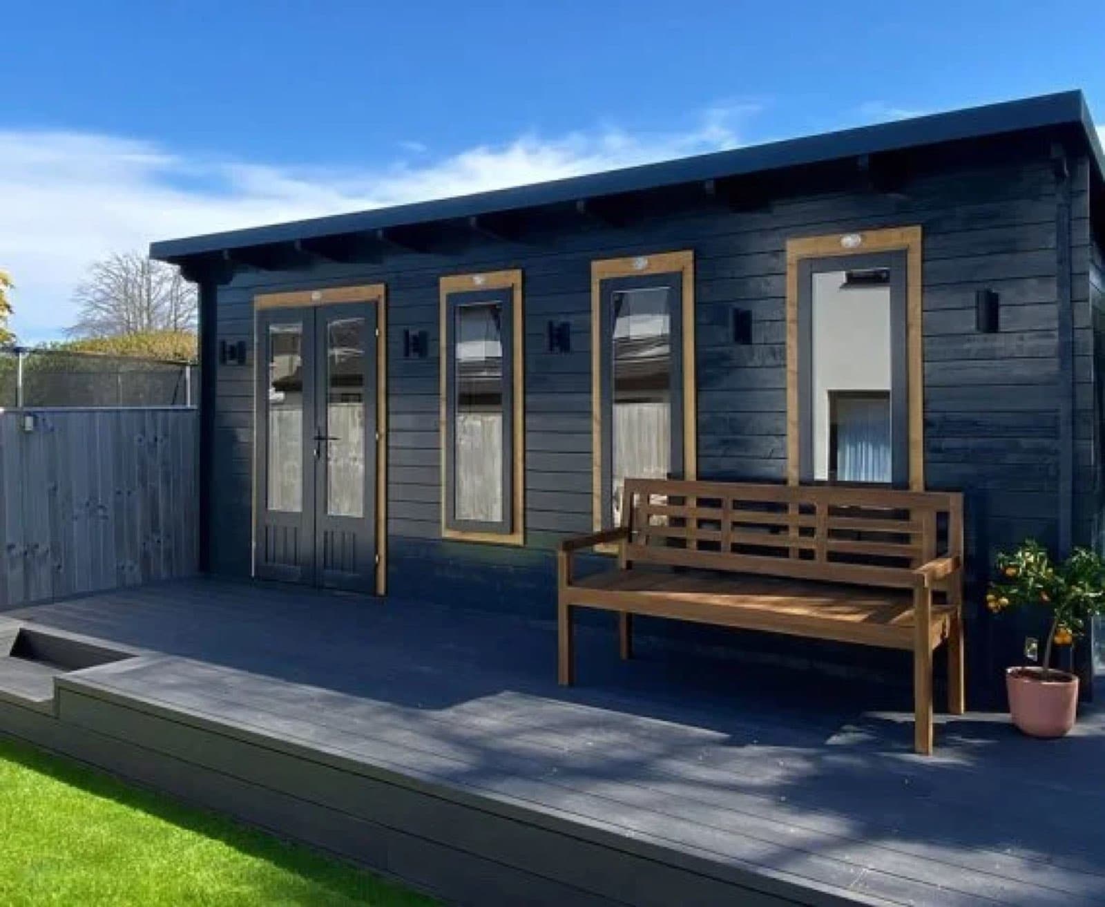 Black painted log cabin with oak-trimmed windows and doors, on a grey composite deck with a timber bench
