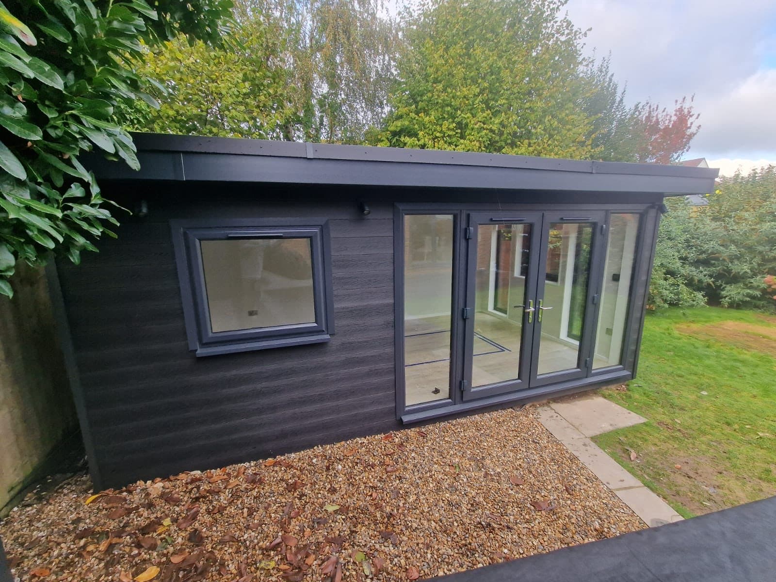 Black composite-clad garden room with grey bifold doors and a large side window, on a gravel base beside a lawn