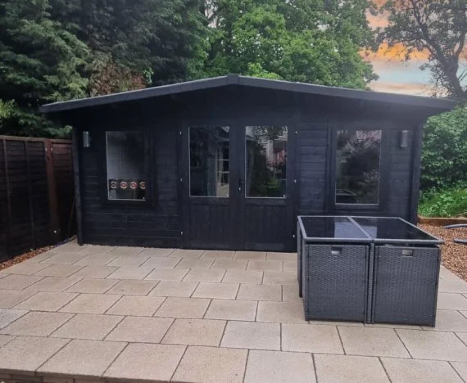 Black painted apex log cabin with glazed doors and windows, on a large paved patio with a rattan dining set