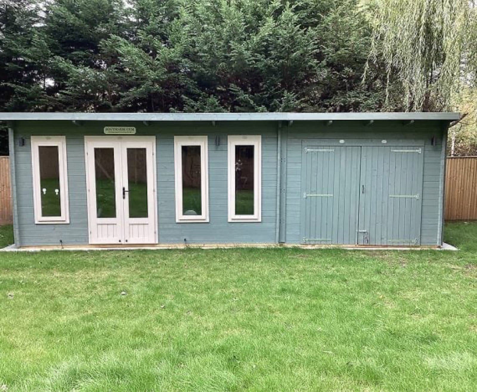 Blue-grey painted pent log cabin with glazed doors, windows and double garage doors at one end, on a lawn backed by conifers