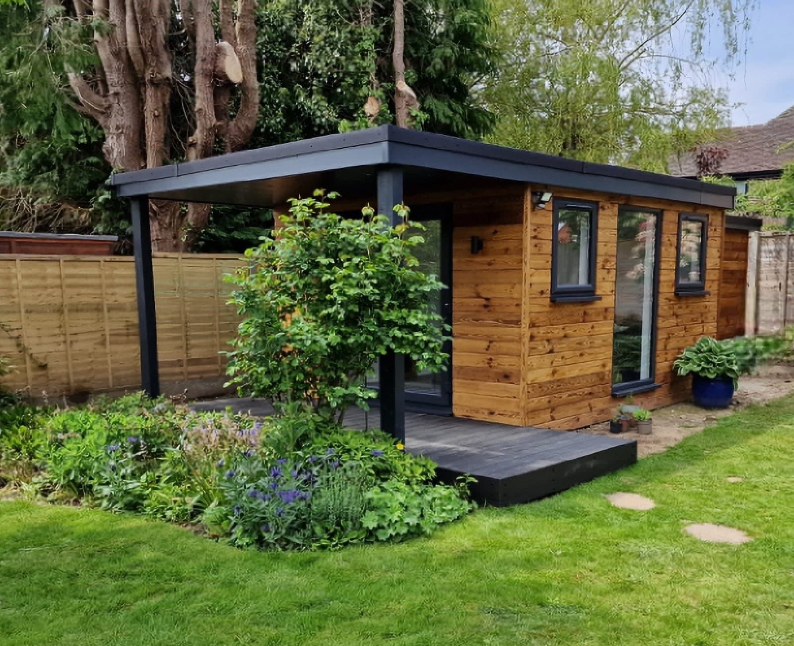 Cedar-clad garden room with an overhanging flat roof forming a canopy over the deck, with cottage garden planting in front