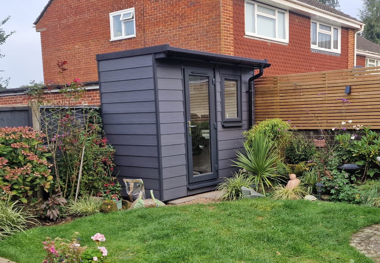 Compact anthracite garden office with a pent roof, a uPVC door and a side window, set into a cottage garden border
