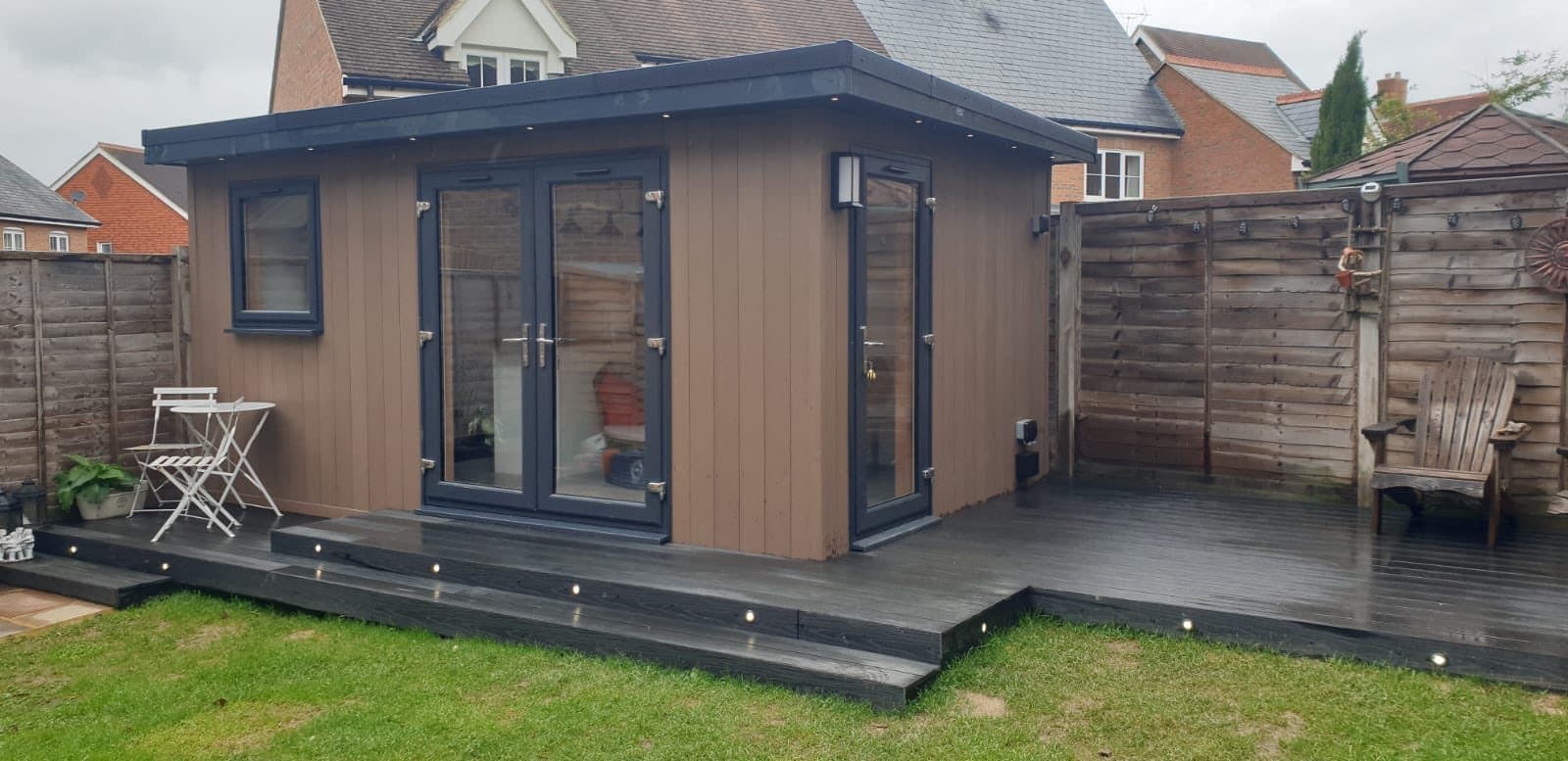 Woodgrain composite-clad garden room with anthracite doors, a composite deck with inset lights and a bistro set to one side