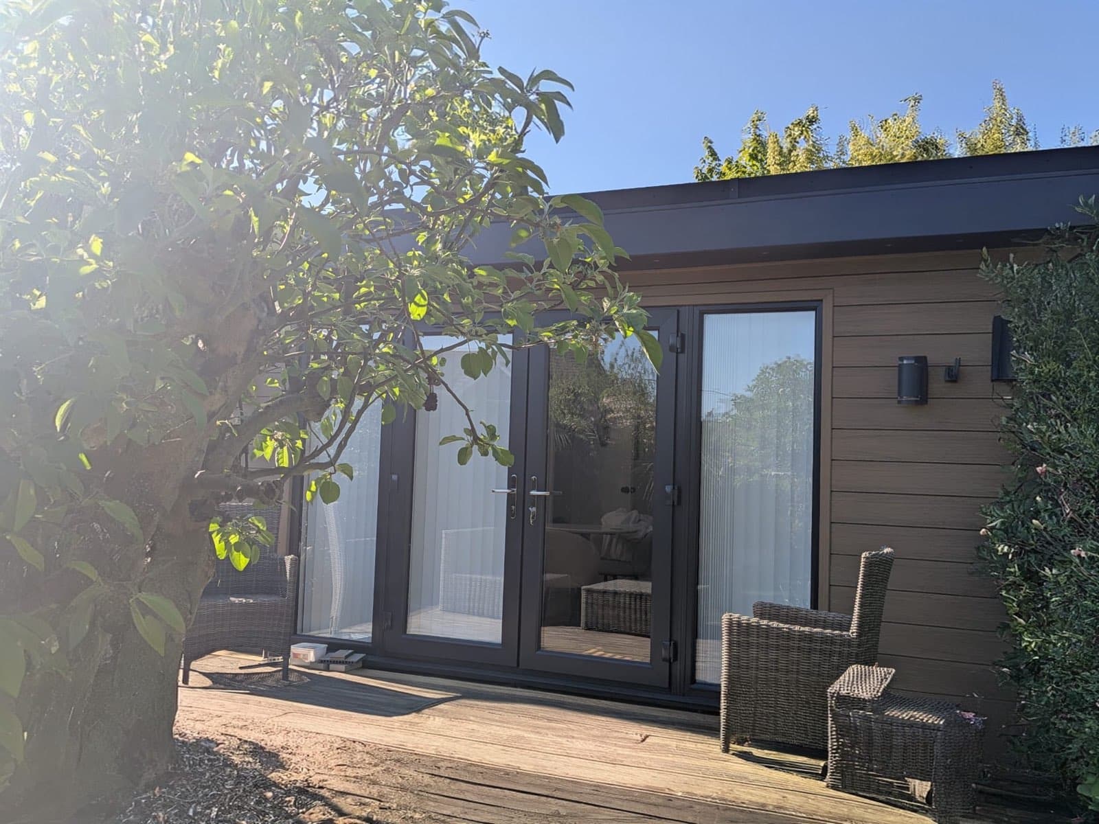 Composite-clad garden room with anthracite French doors and a timber deck with rattan chairs, under an apple tree