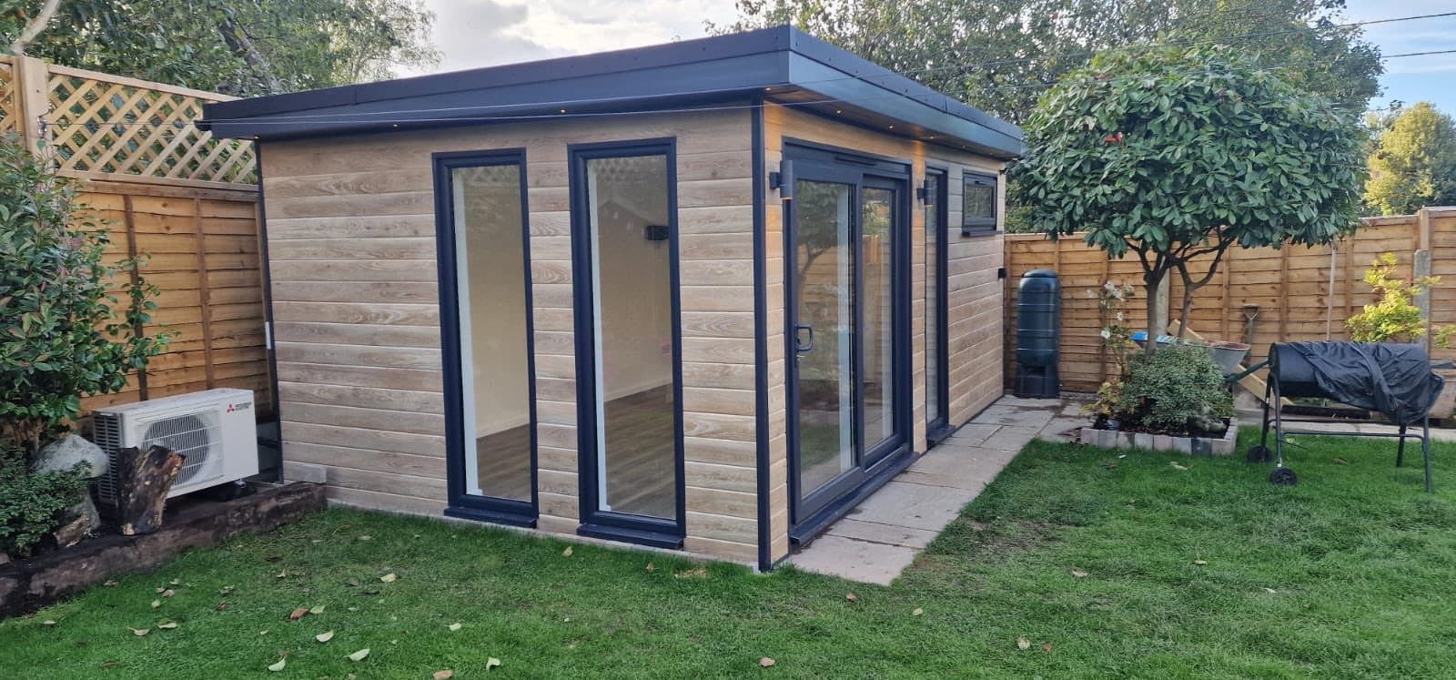 Woodgrain composite-clad garden room with anthracite sliding doors and an air source heat pump, on a lawn