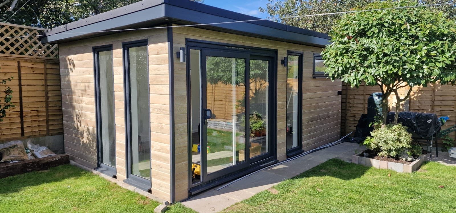 Garden room clad in woodgrain composite with wide anthracite sliding doors, opening onto a lawn
