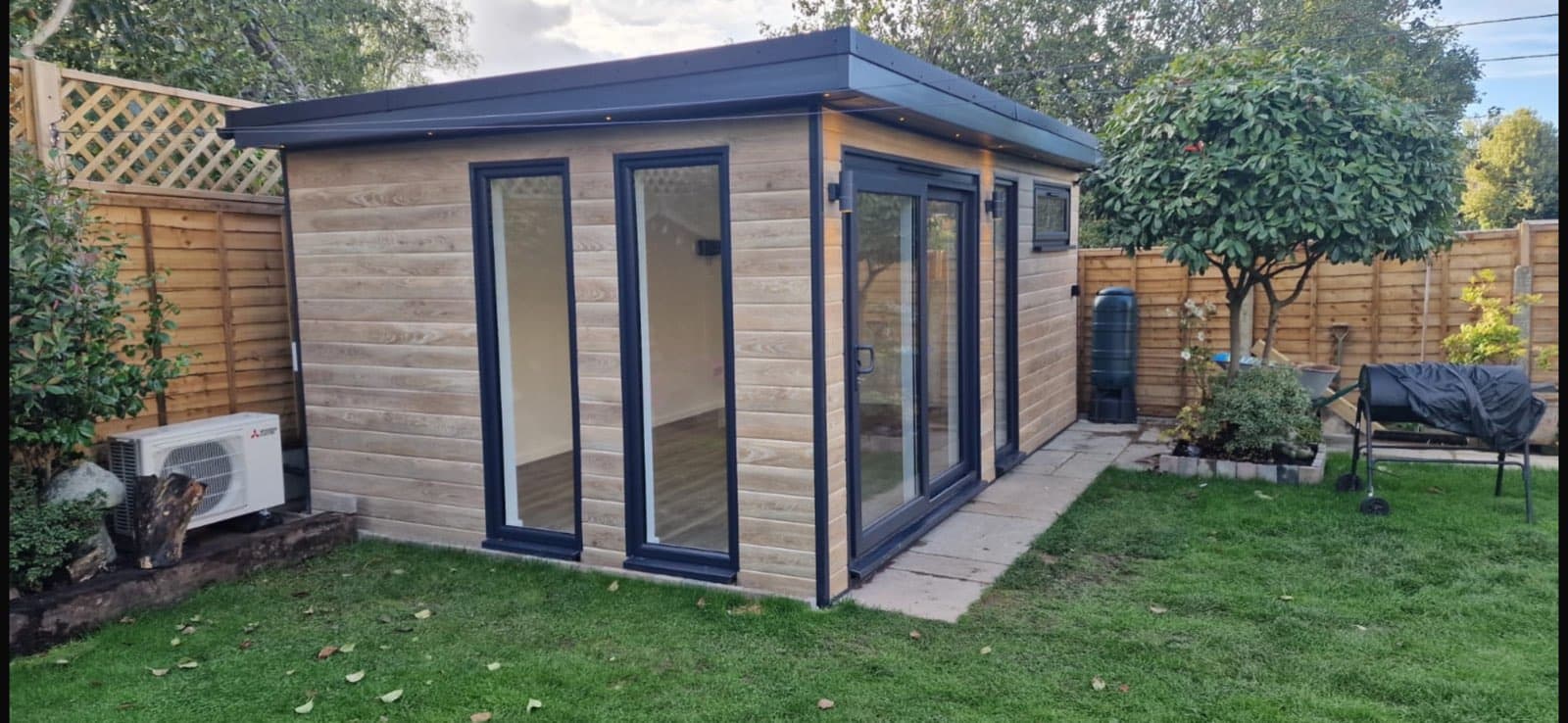 Garden room clad in woodgrain composite with anthracite sliding doors, an air source heat pump at one end and a lawn in front