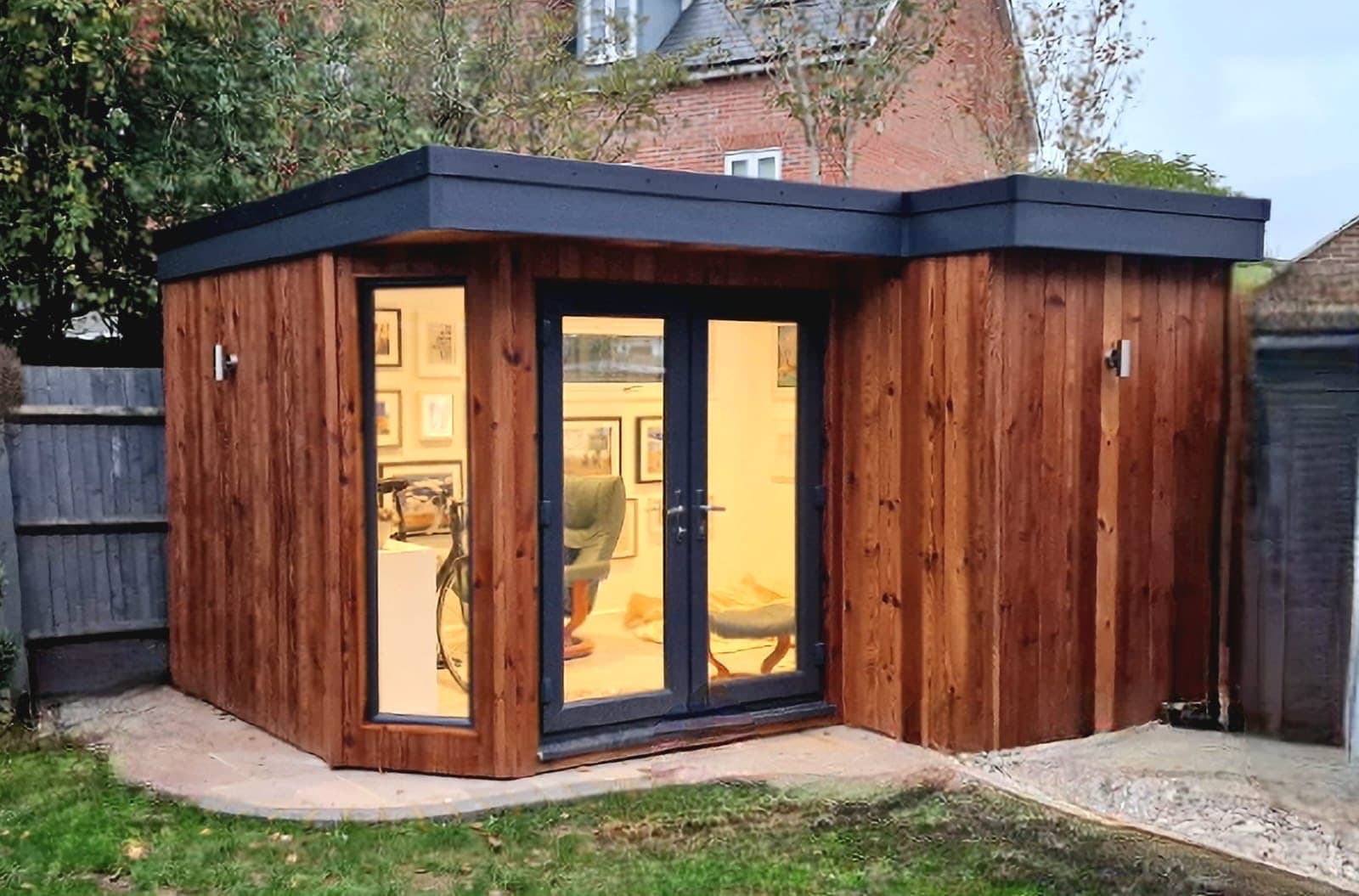 Dark-stained corner garden room with a flat roof and French doors, lit inside to show framed artwork, beside a brick house