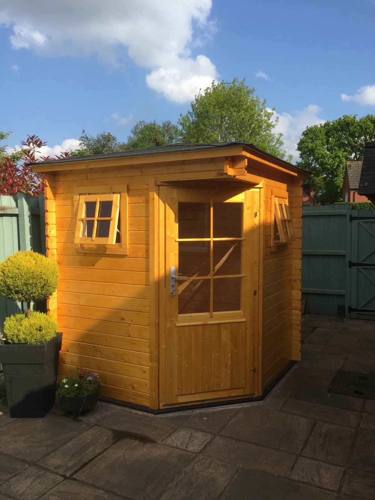 Honey-stained corner summerhouse with a pent roof and a single glazed door, on a paved patio with clipped topiary in planters