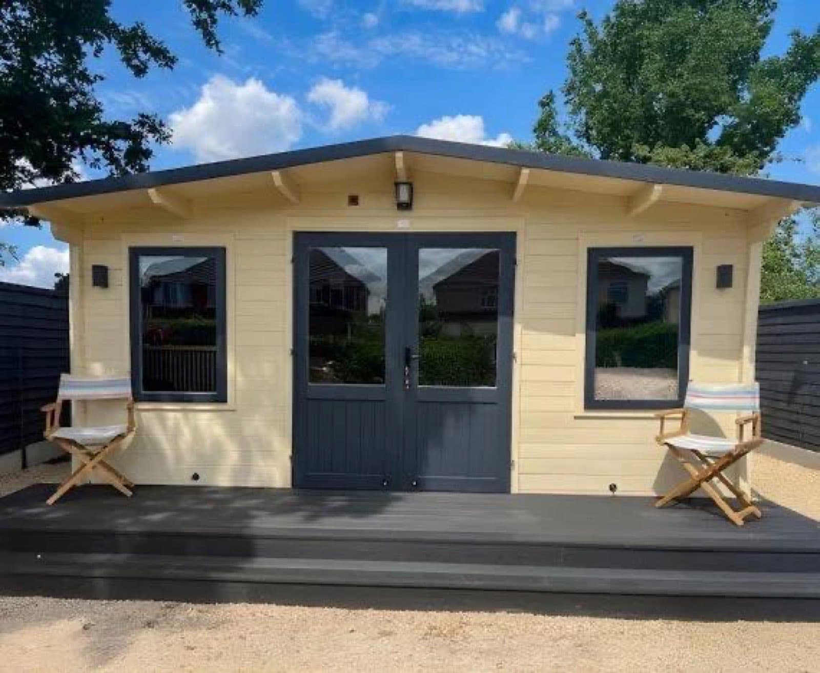Cream painted log cabin with anthracite doors and windows, on a grey composite deck with two directors chairs