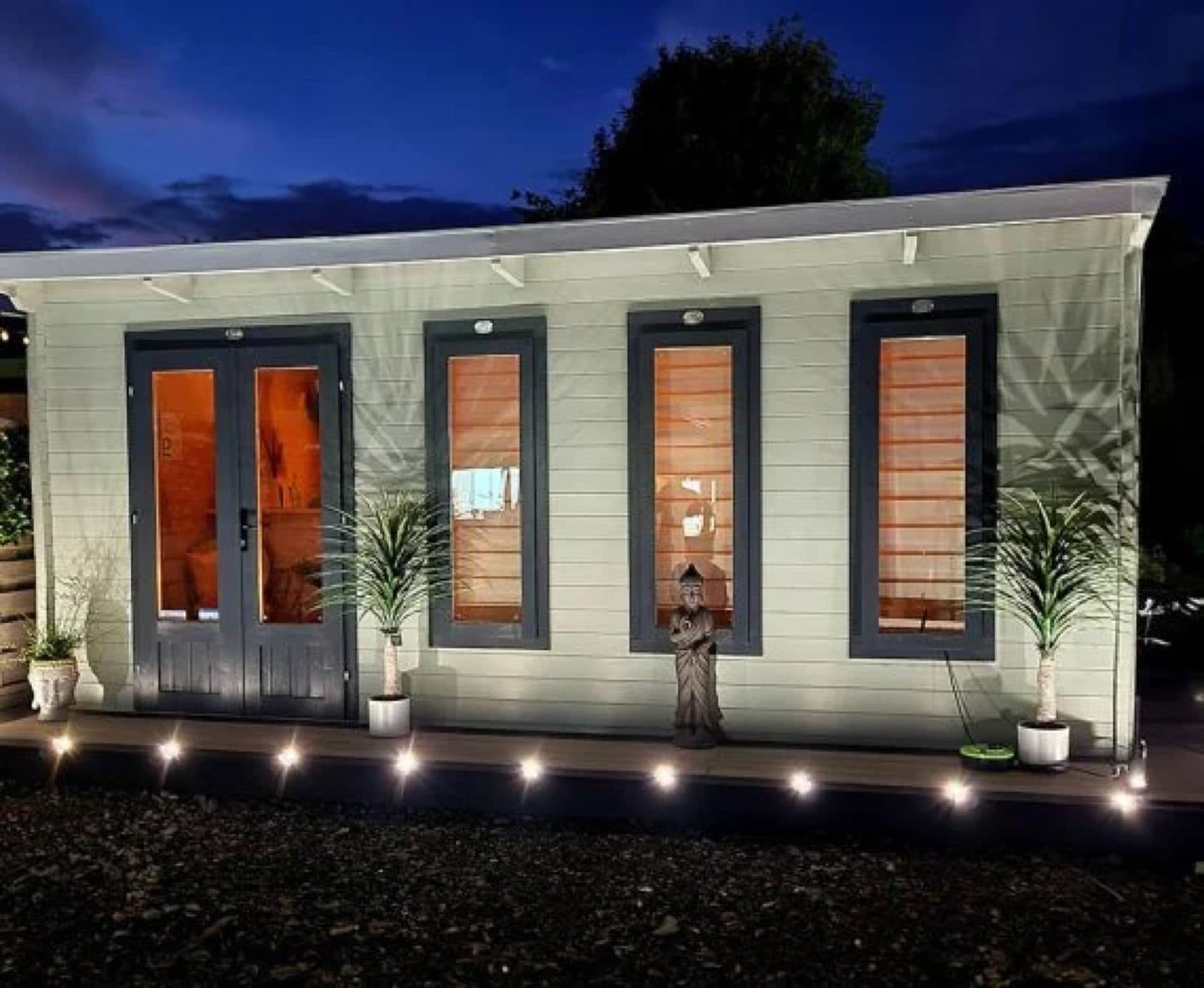 Cream painted log cabin lit from inside at dusk, with deck lights along the front and potted palms either side