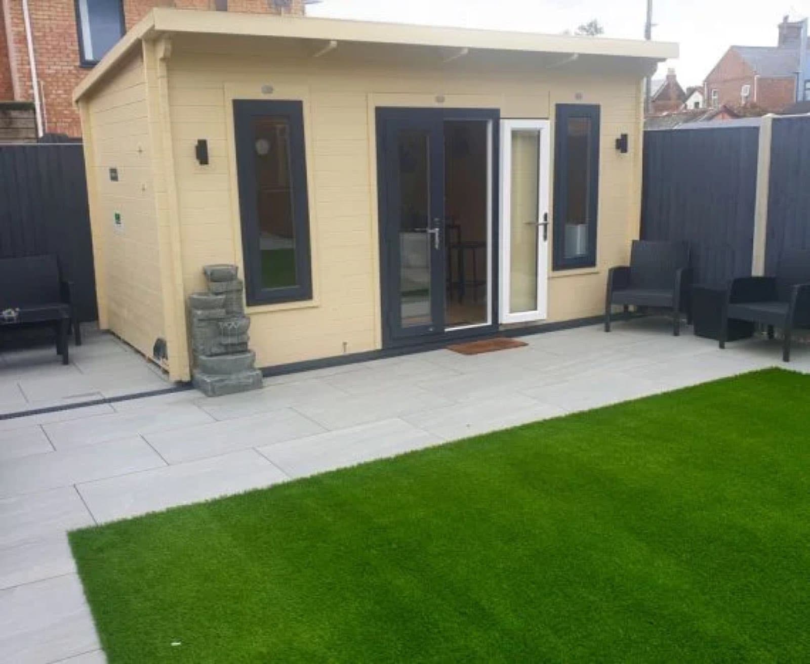 Cream painted pent log cabin with anthracite windows and a white door, on porcelain paving beside artificial lawn