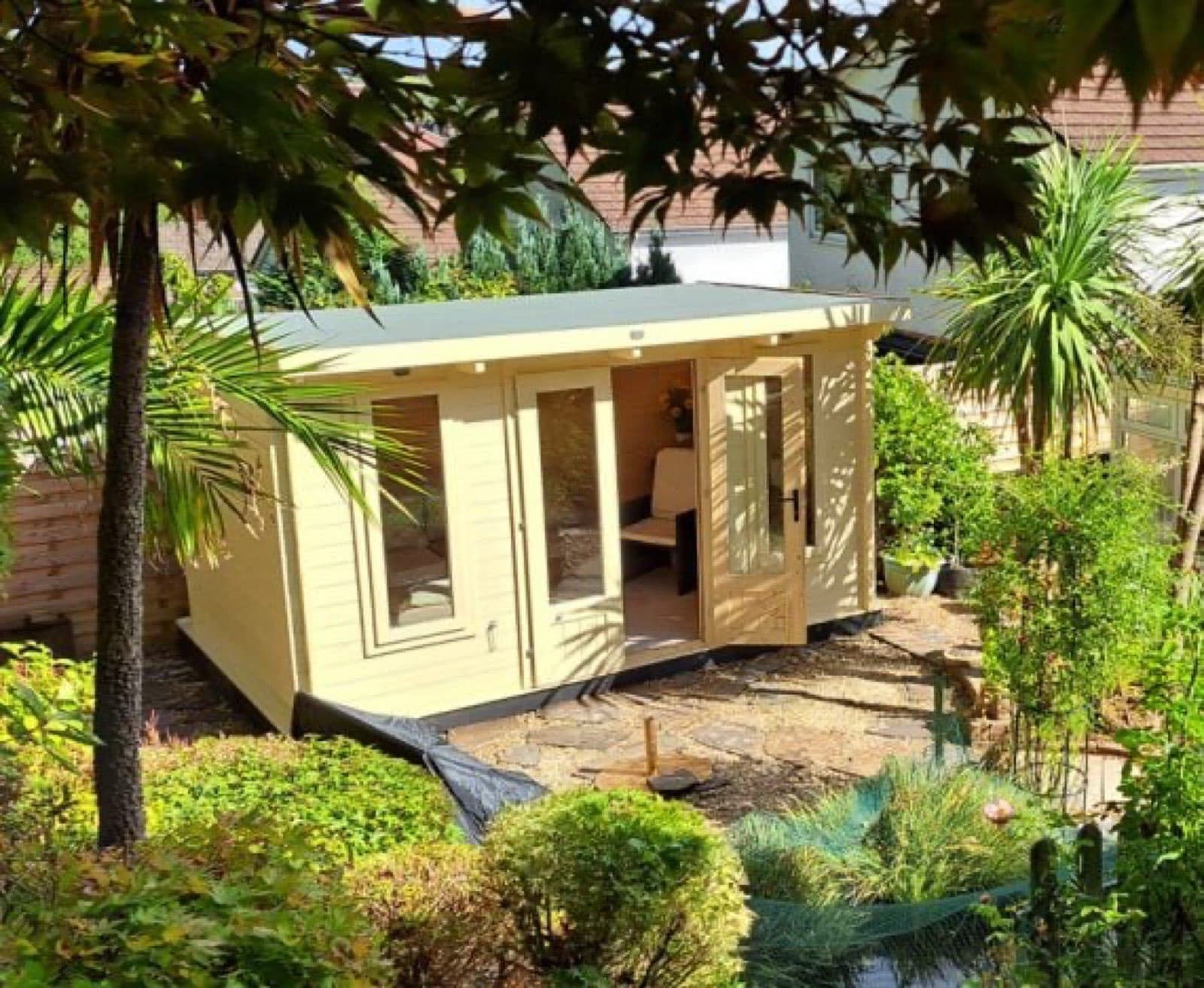 Cream painted pent log cabin with open doors and a desk inside, surrounded by palms and tropical planting