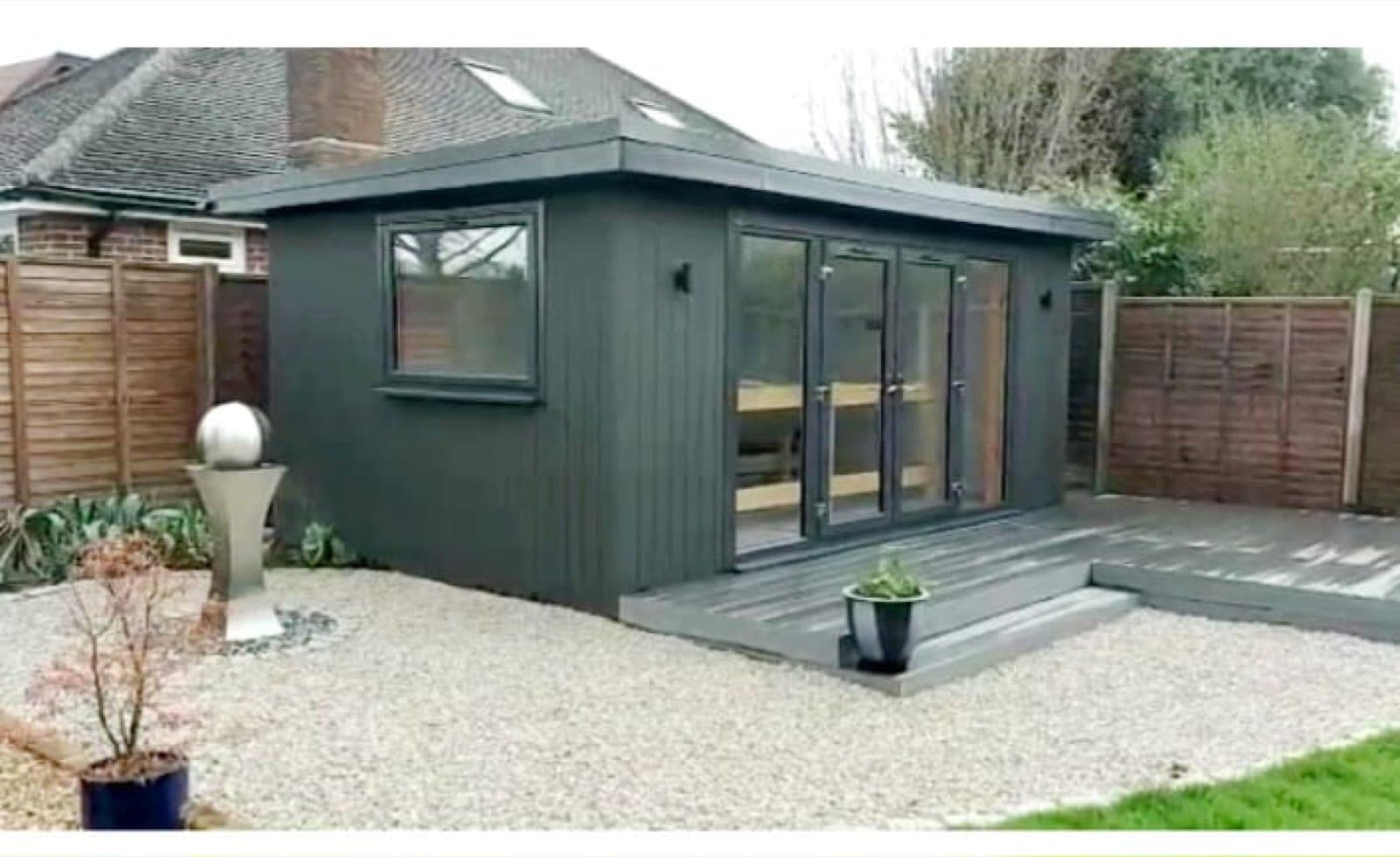 Dark grey composite-clad garden room with bifold doors, on a gravel base with composite decking in front of a bungalow
