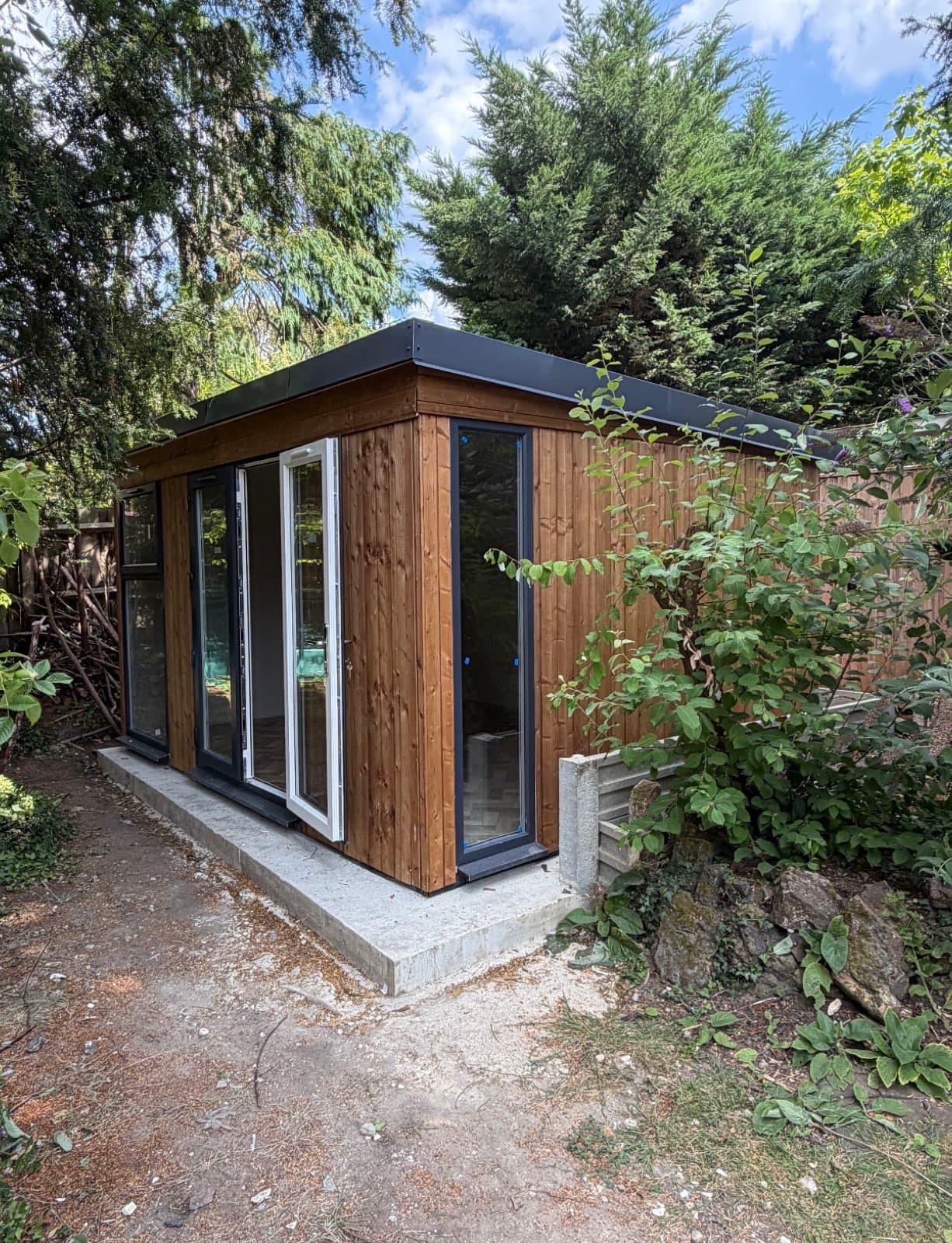 Dark-stained timber garden room with white uPVC French doors, on a concrete base at the edge of woodland