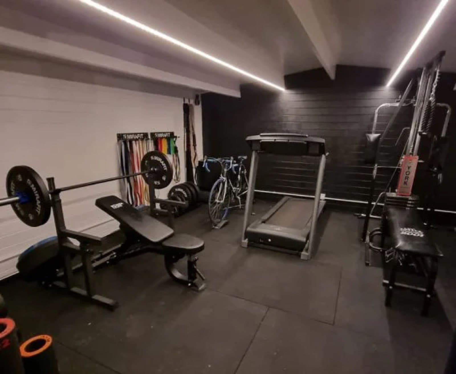 Inside a garden gym with a treadmill, a weights bench and barbell, a rack of weight plates, rubber flooring and LED strip lighting