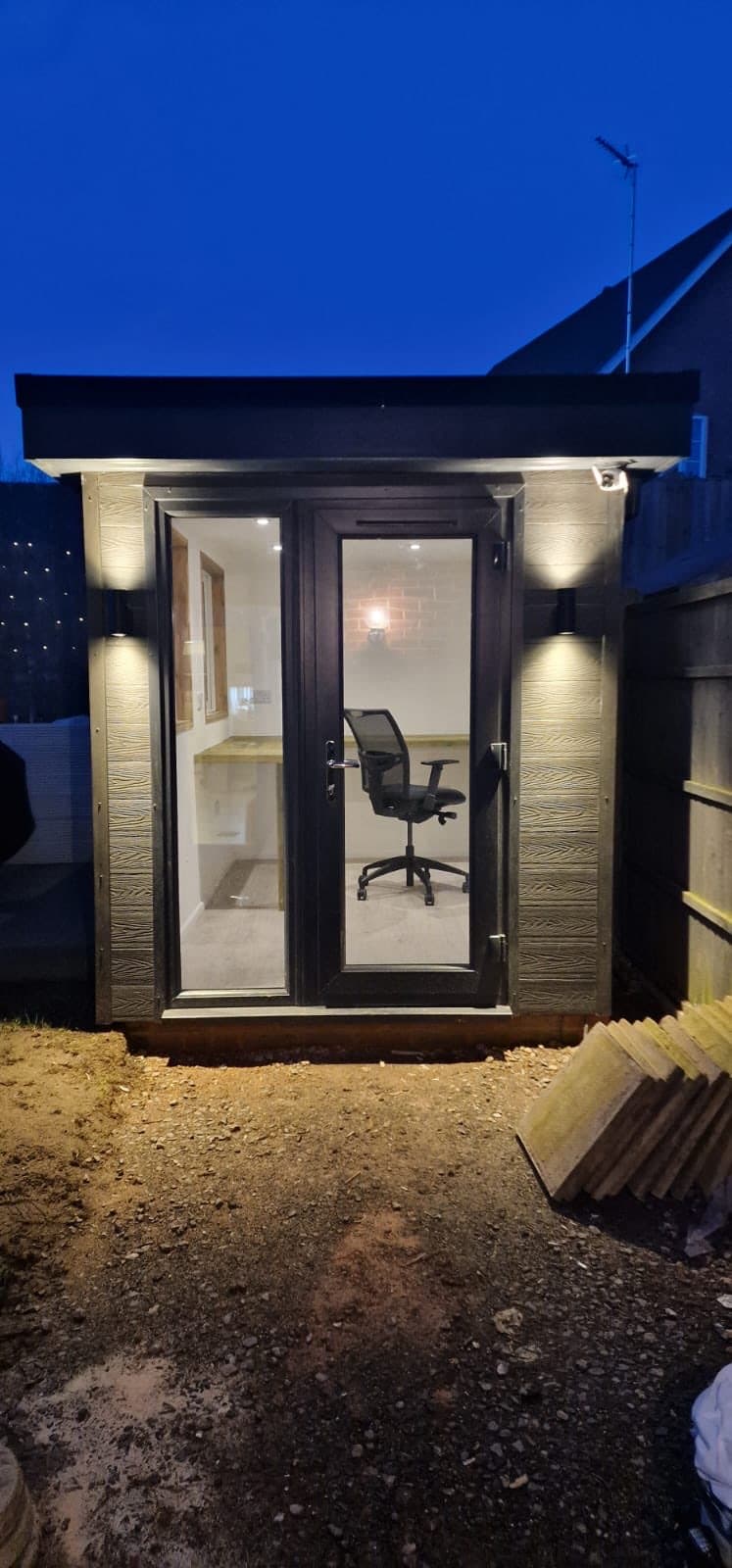 Compact garden office lit at night with a uPVC door and a desk chair visible inside, with groundworks still in progress around it