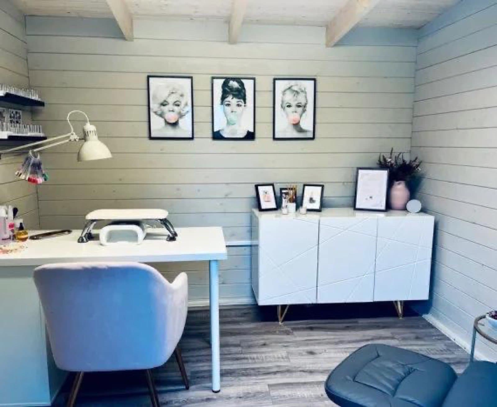 Inside a pale painted garden room fitted out as a beauty salon, with a treatment desk, lamp, framed prints and a white sideboard