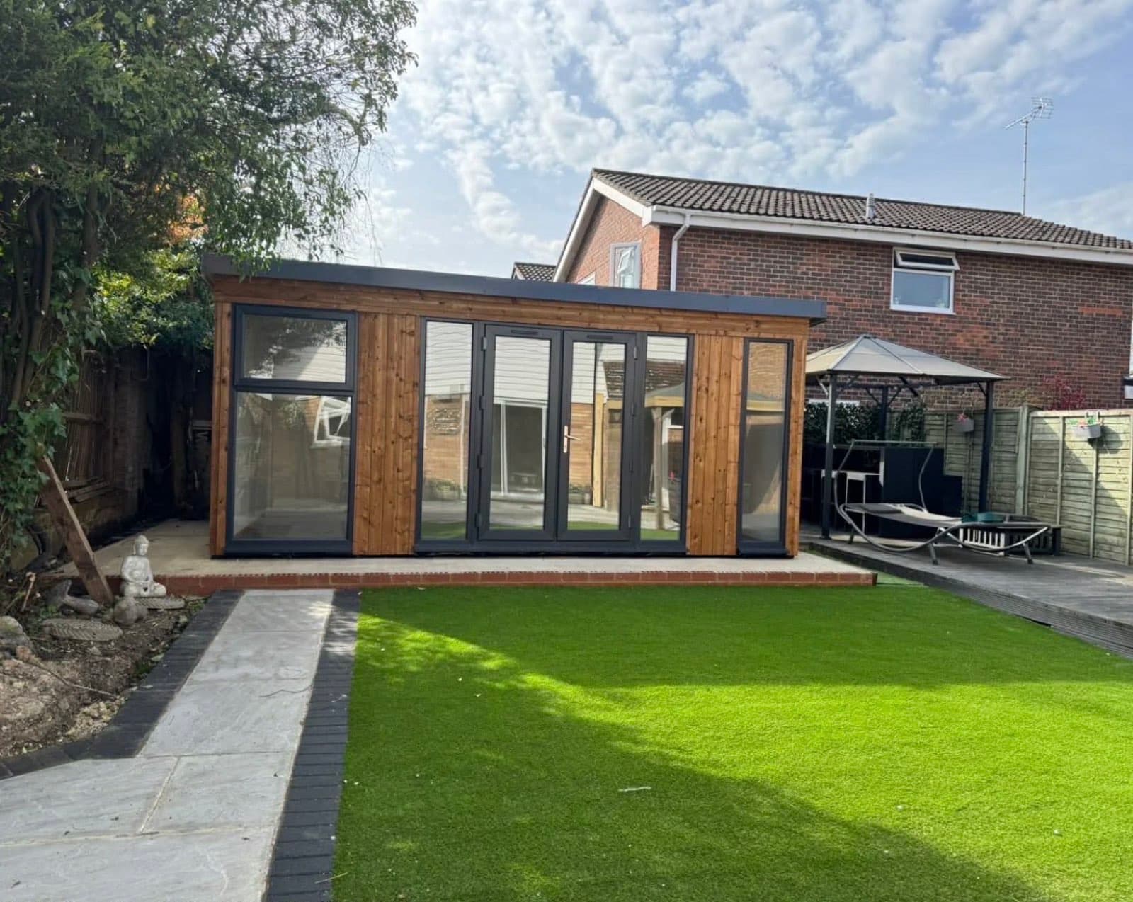 Timber and glass garden room with anthracite bifold doors on a raised deck, with artificial lawn in front and a house behind