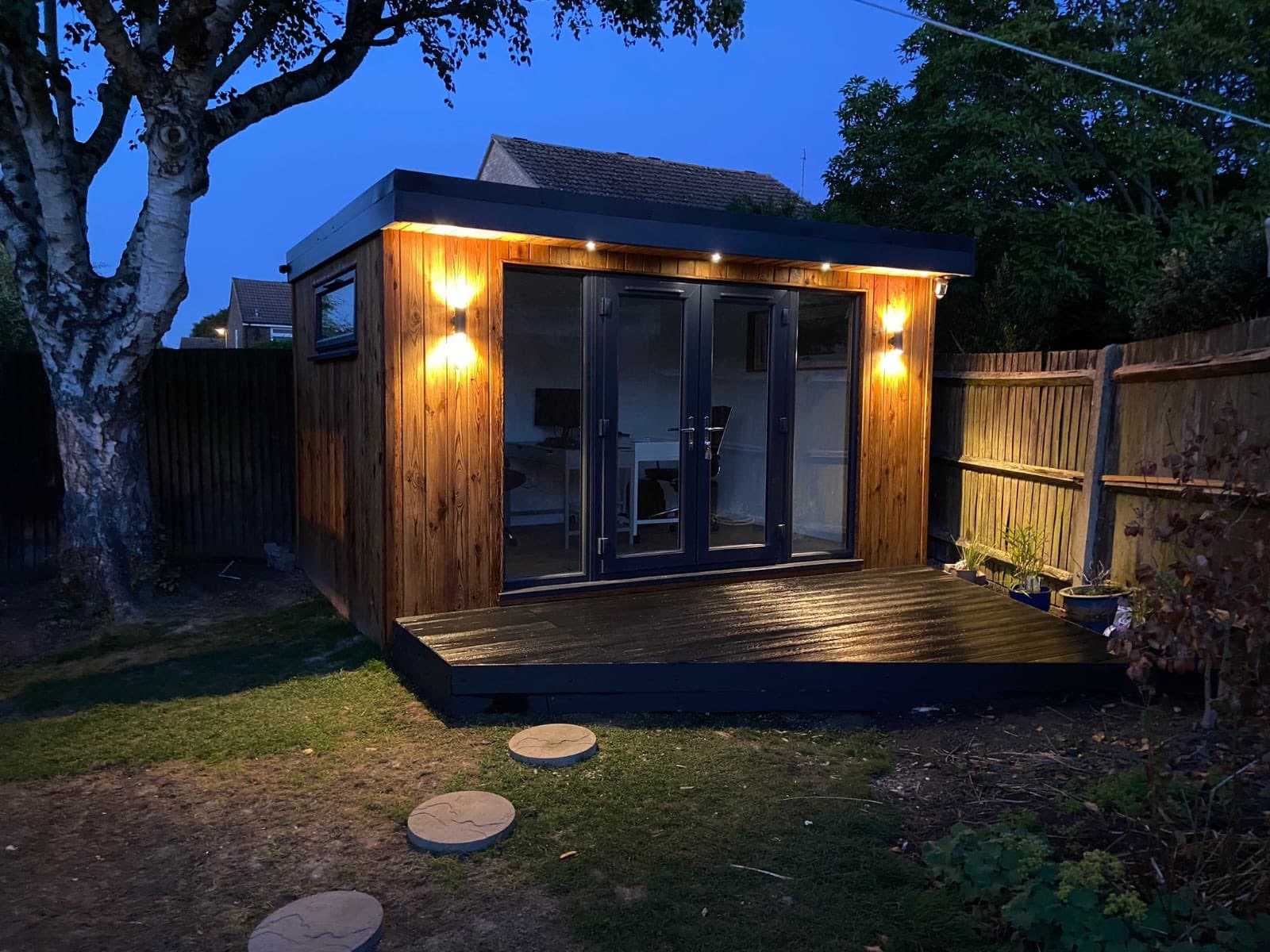 Timber garden room lit at dusk with anthracite French doors, a raised deck in front and a desk visible inside