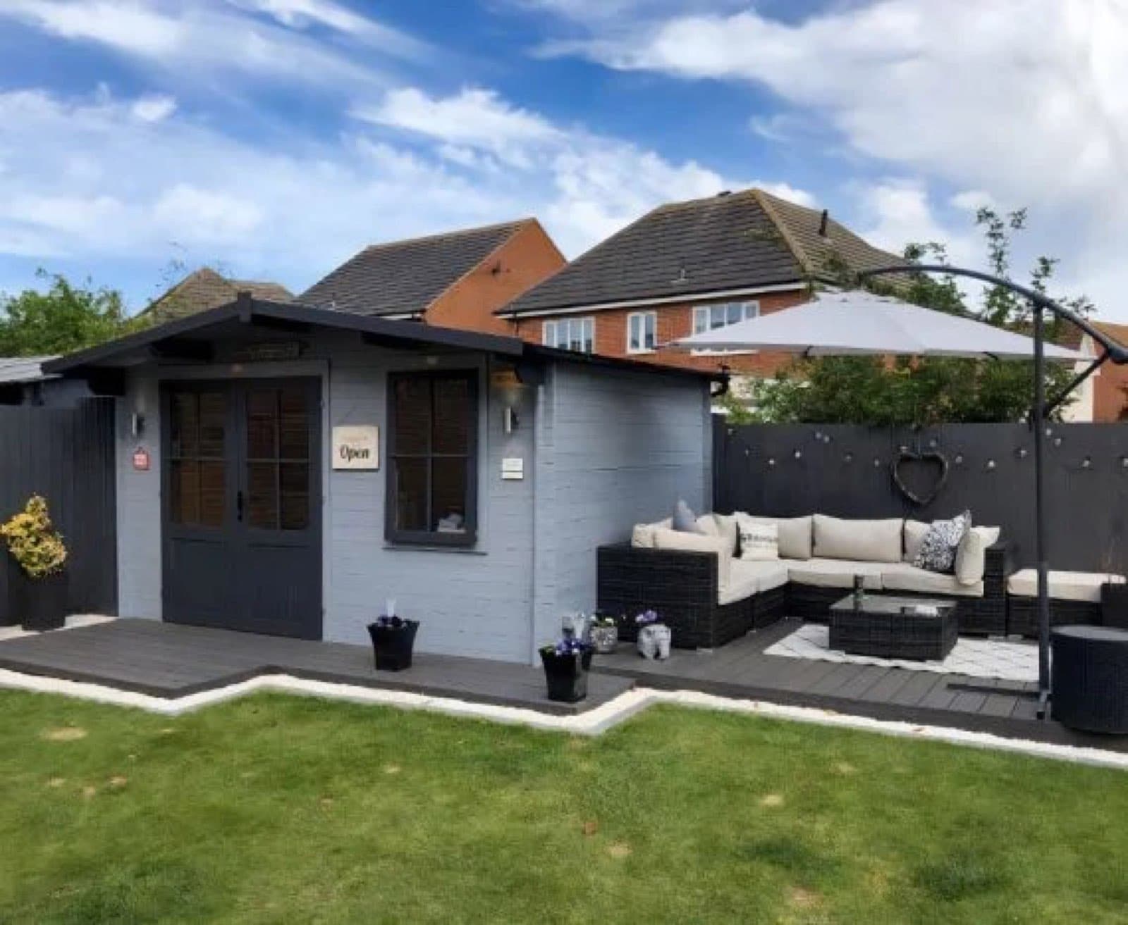 Grey painted apex summerhouse with an Open sign, a composite deck, a rattan corner sofa and a parasol