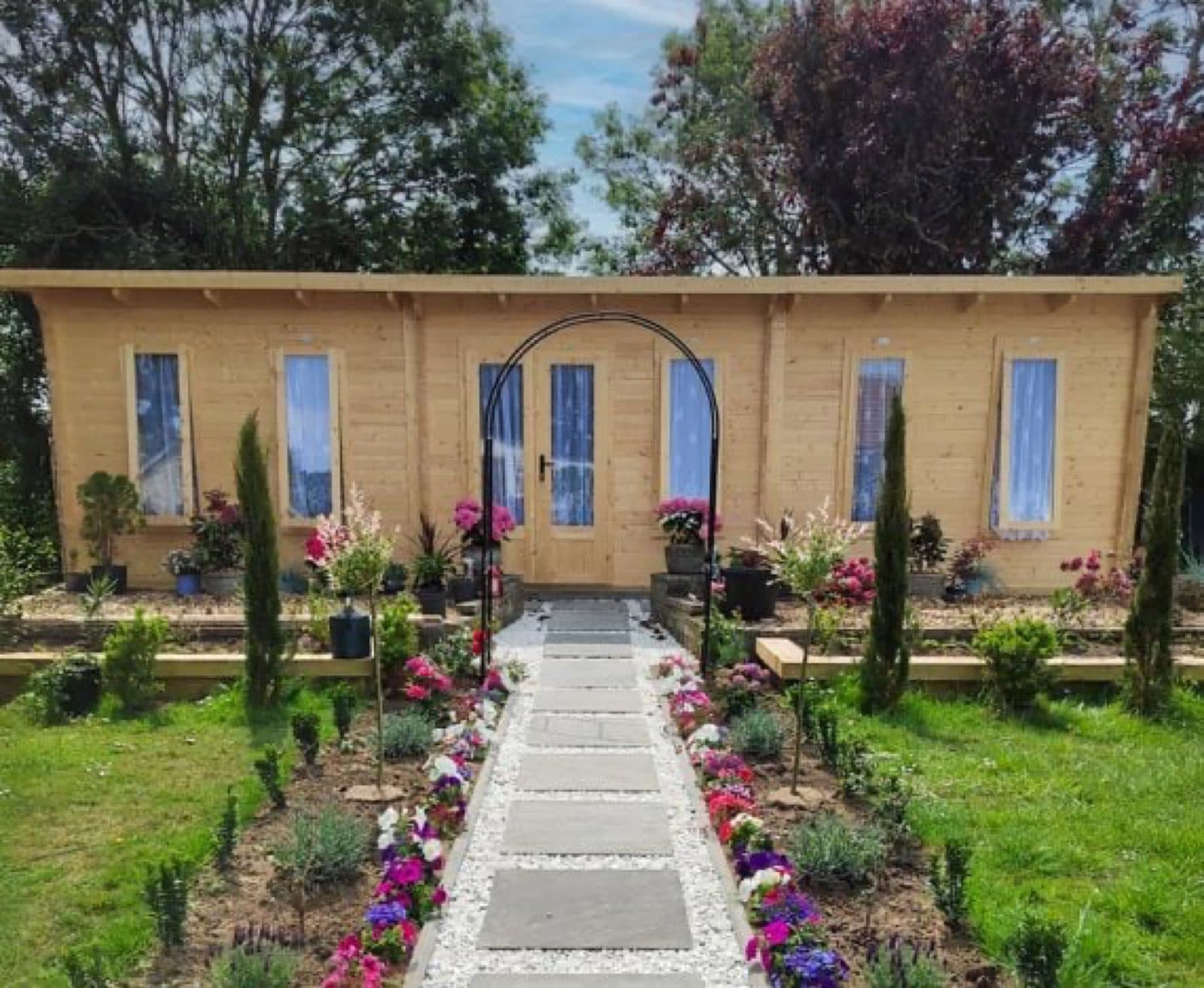 Honey log cabin with glazed double doors and curtains, in a formal garden with flower beds and a path through a rose arch