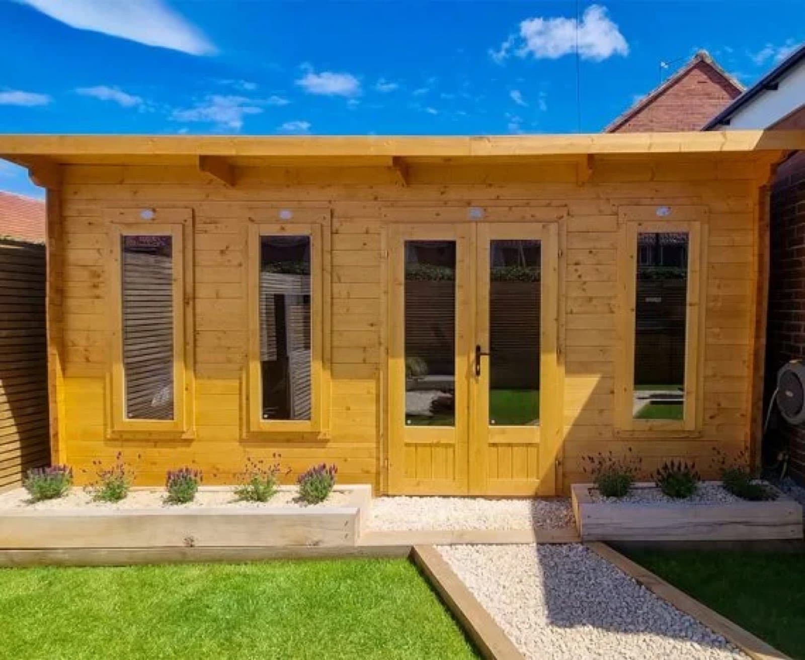 Honey-stained pent log cabin with glazed double doors and two windows, with lavender in raised beds and a gravel path in front