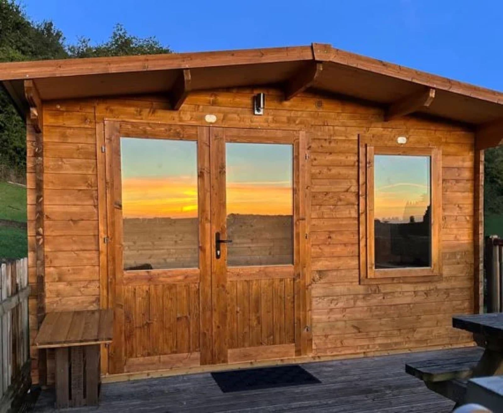 Honey log cabin at sunset with the sky reflected in its glazed doors, with decking and a picnic bench in front