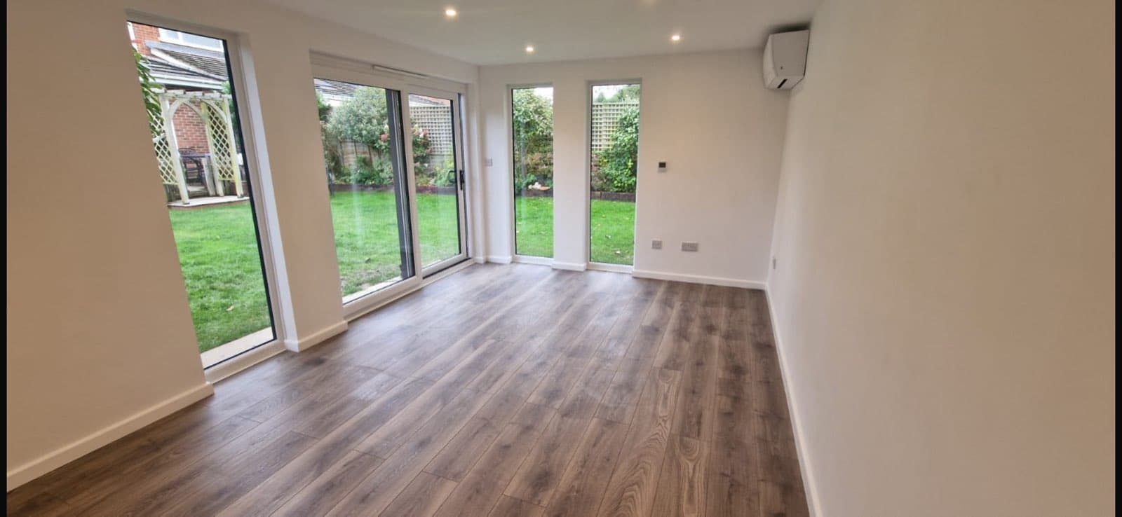 Inside an empty insulated garden room with three sets of glazed doors onto a lawn, dark laminate flooring and a wall-mounted air conditioning unit