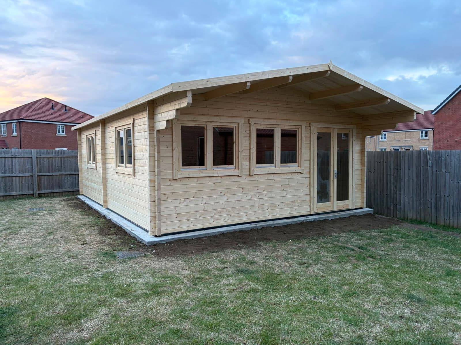 Large untreated apex log cabin with a run of windows and glazed doors, on a lawn behind a new-build house