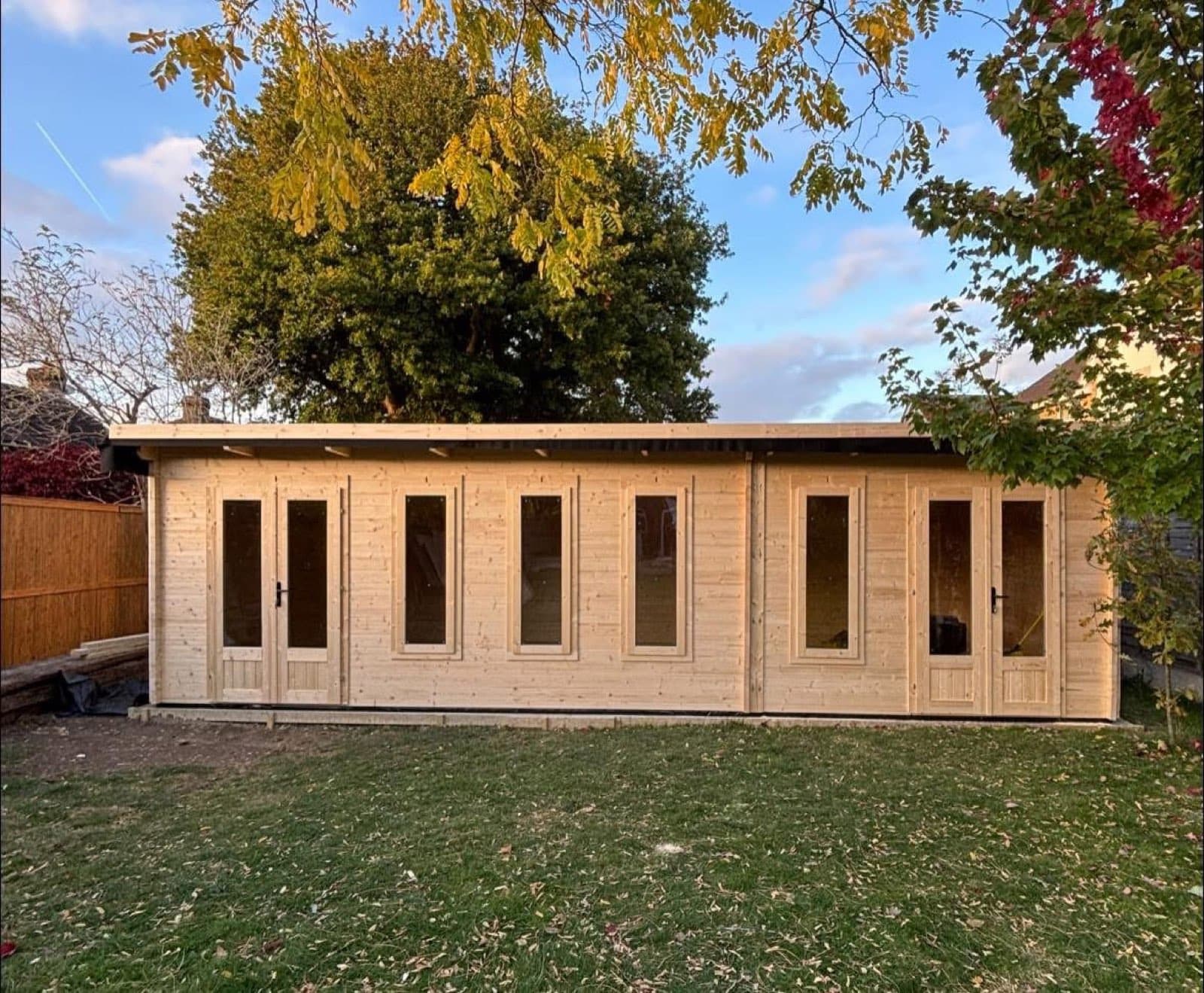Large untreated multi-room pent log cabin with four sets of glazed doors across the front, on a lawn under an autumn tree
