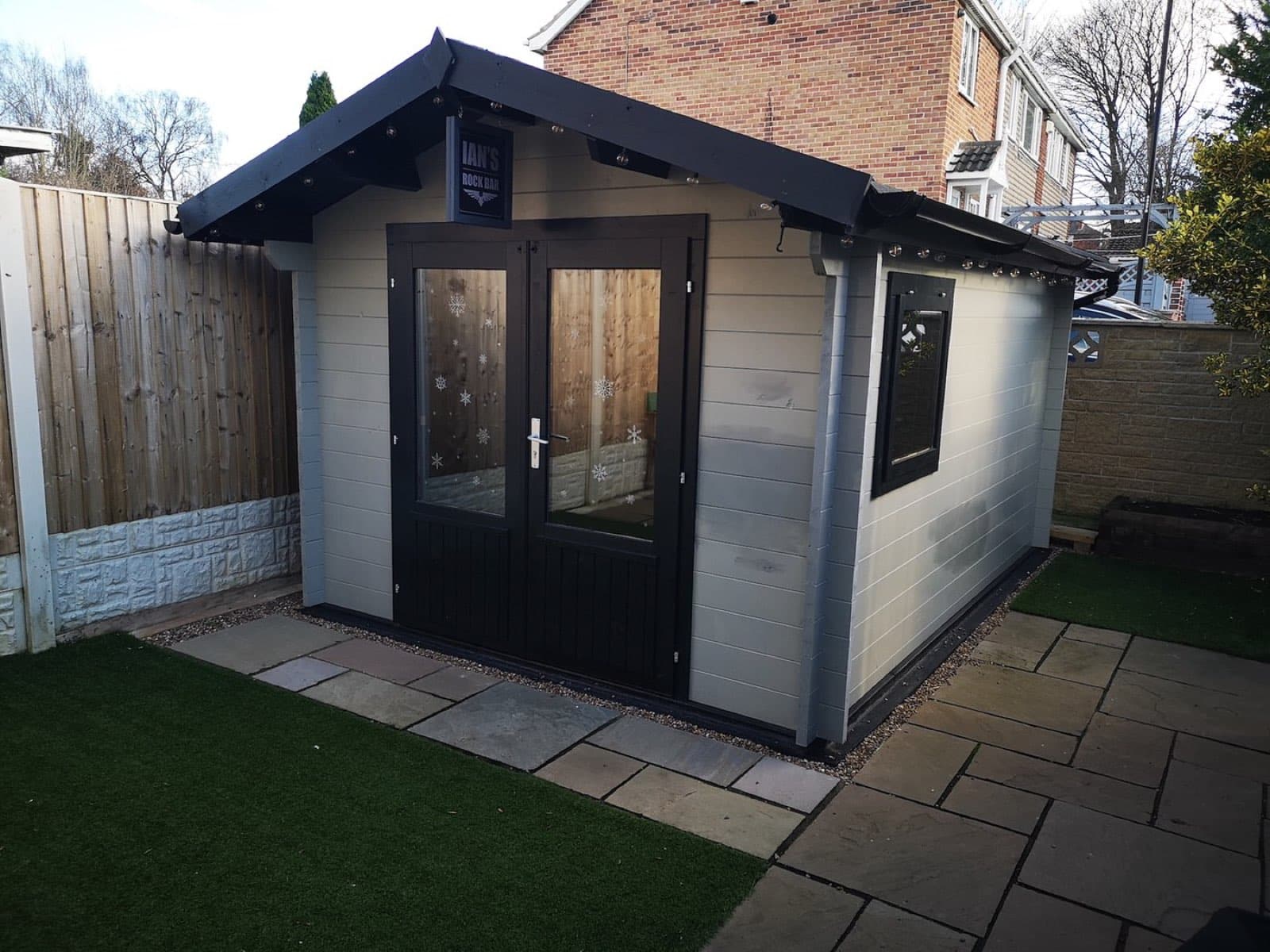 Light grey log cabin with a black apex roof, black glazed double doors and festoon lights, on sandstone paving beside artificial lawn