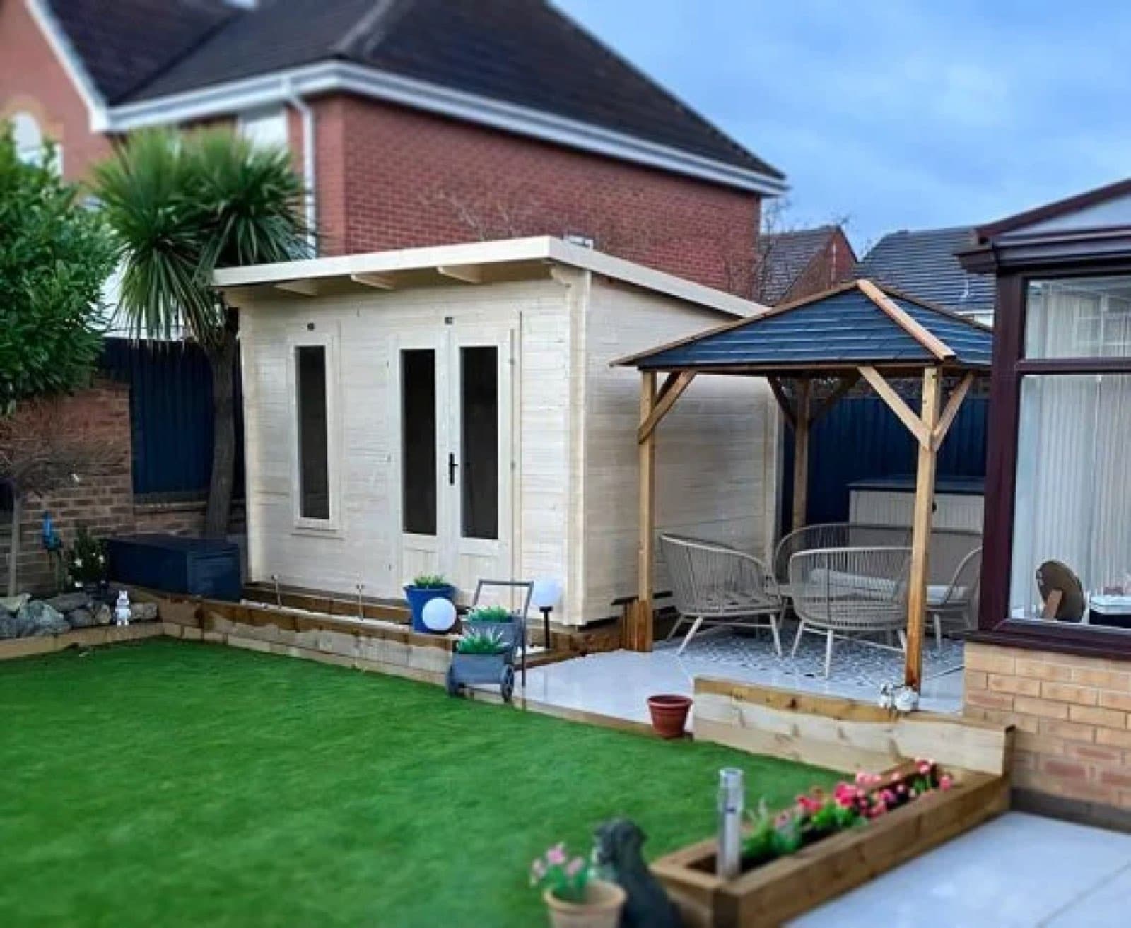 Pale untreated pent log cabin next to a timber gazebo, with a lawn and patio in front