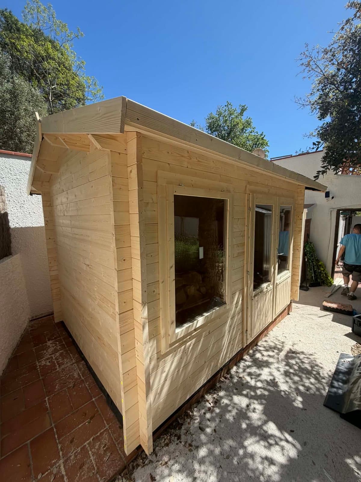 Untreated pent log cabin in a walled courtyard, with a large picture window and terracotta tiles underfoot