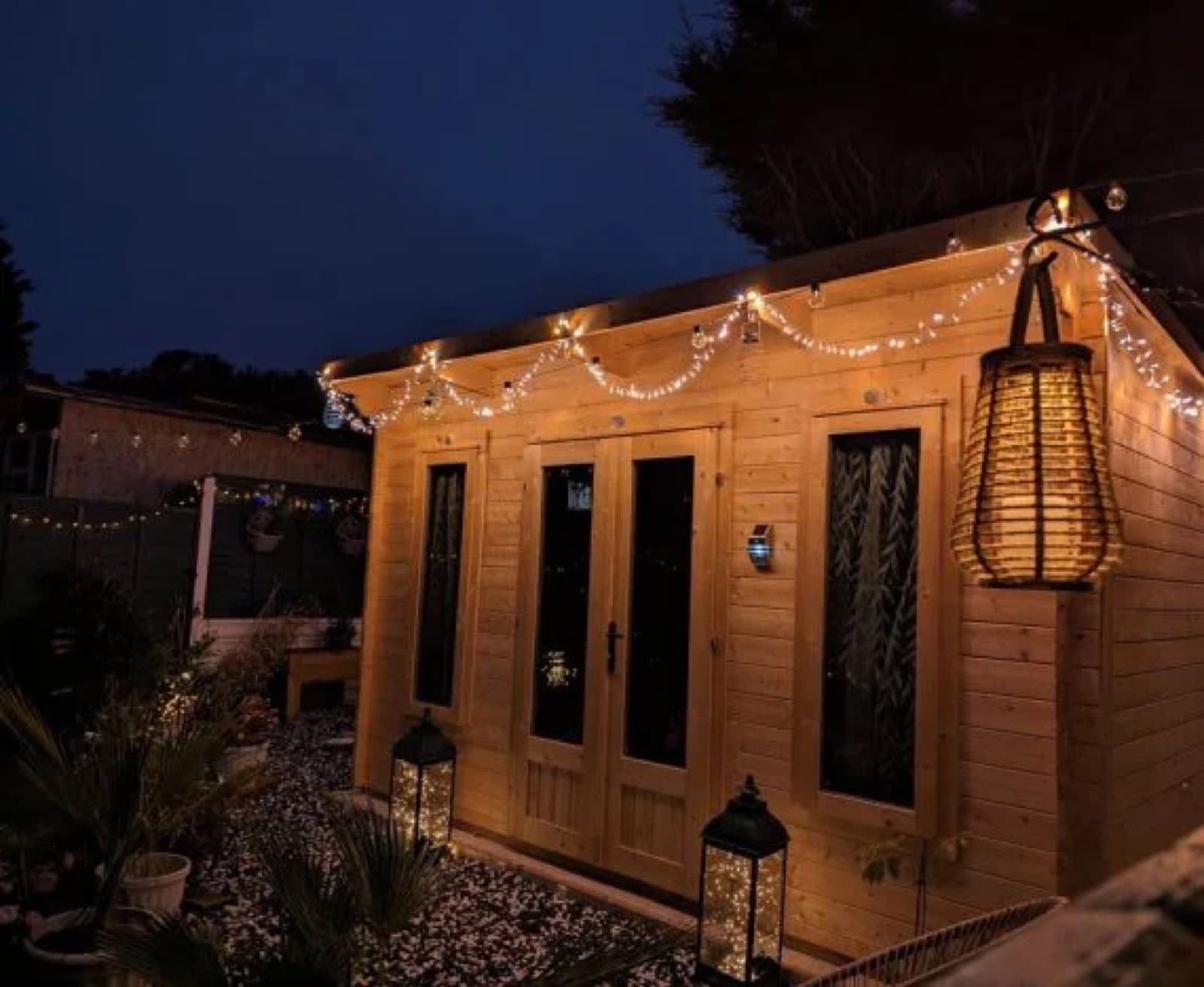 Honey log cabin at night strung with fairy lights, with lanterns on the gravel and glazed double doors