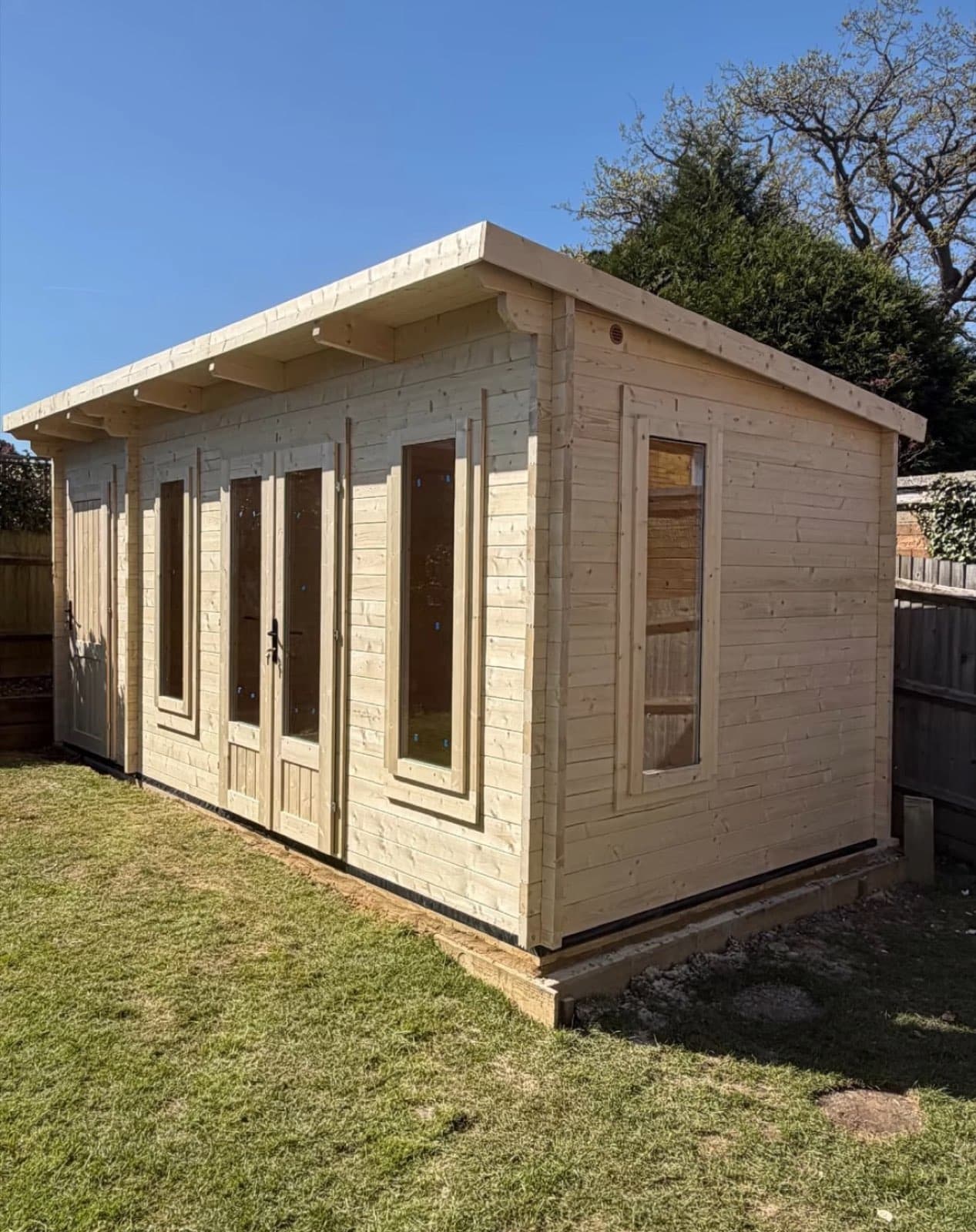 Untreated pent-roof log cabin with full-height windows either side of glazed double doors, on a lawn