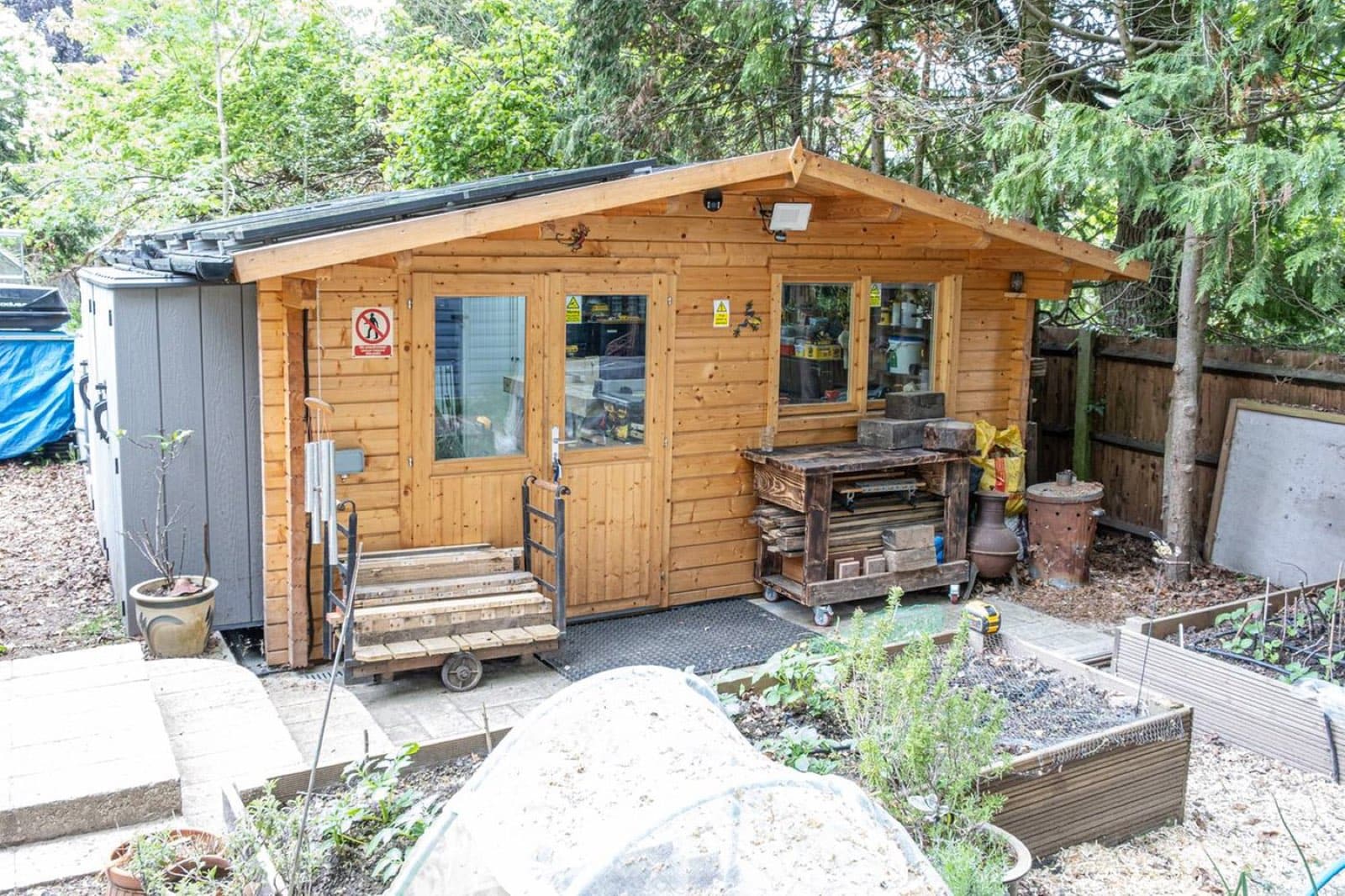 Honey-coloured log cabin used as a garden workshop, with a potting bench outside and raised vegetable beds in front