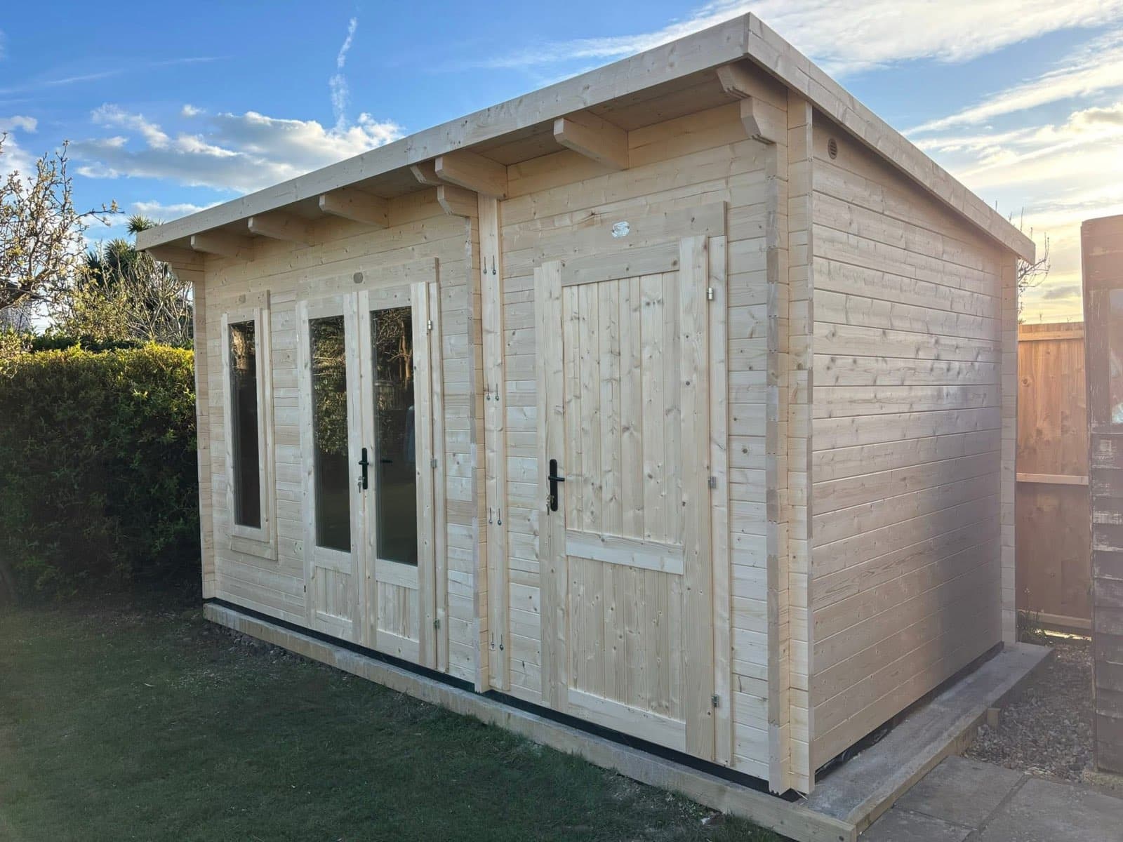 Untreated pent log cabin with glazed double doors, a window and a separate store door, on a lawn at sunset