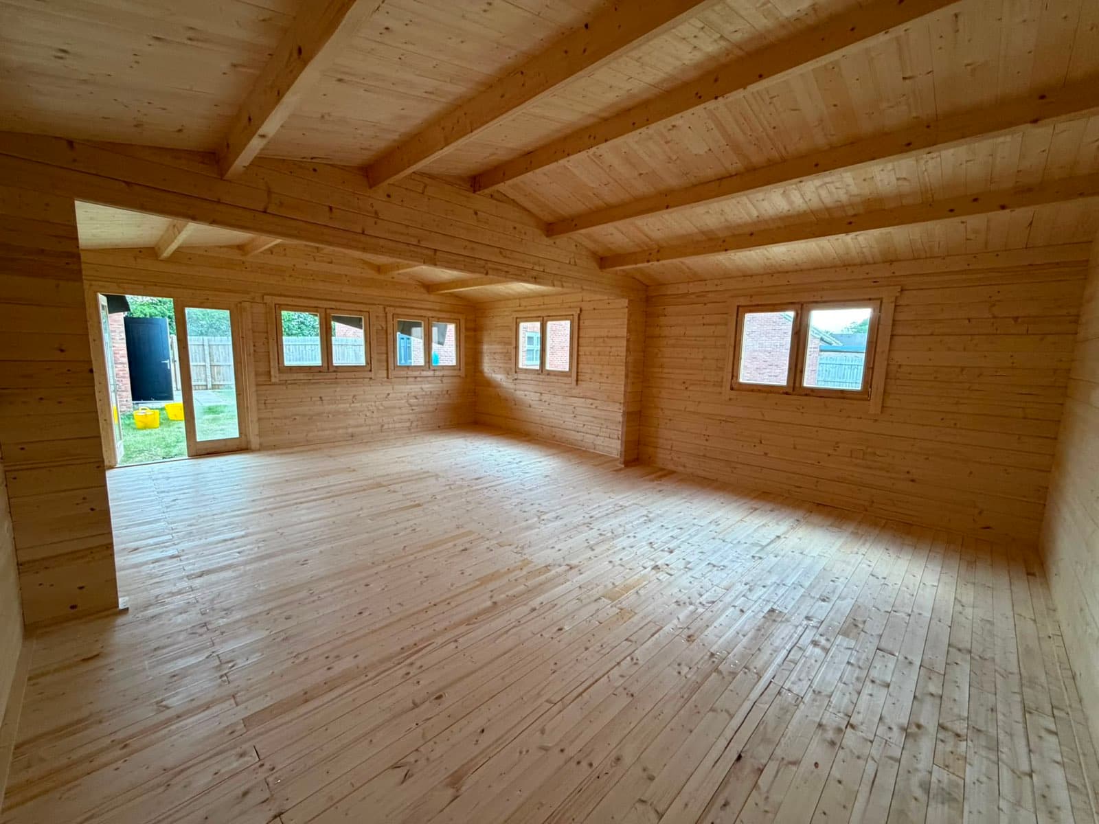 Inside a large untreated log cabin with exposed roof purlins, a timber floor and windows along two walls