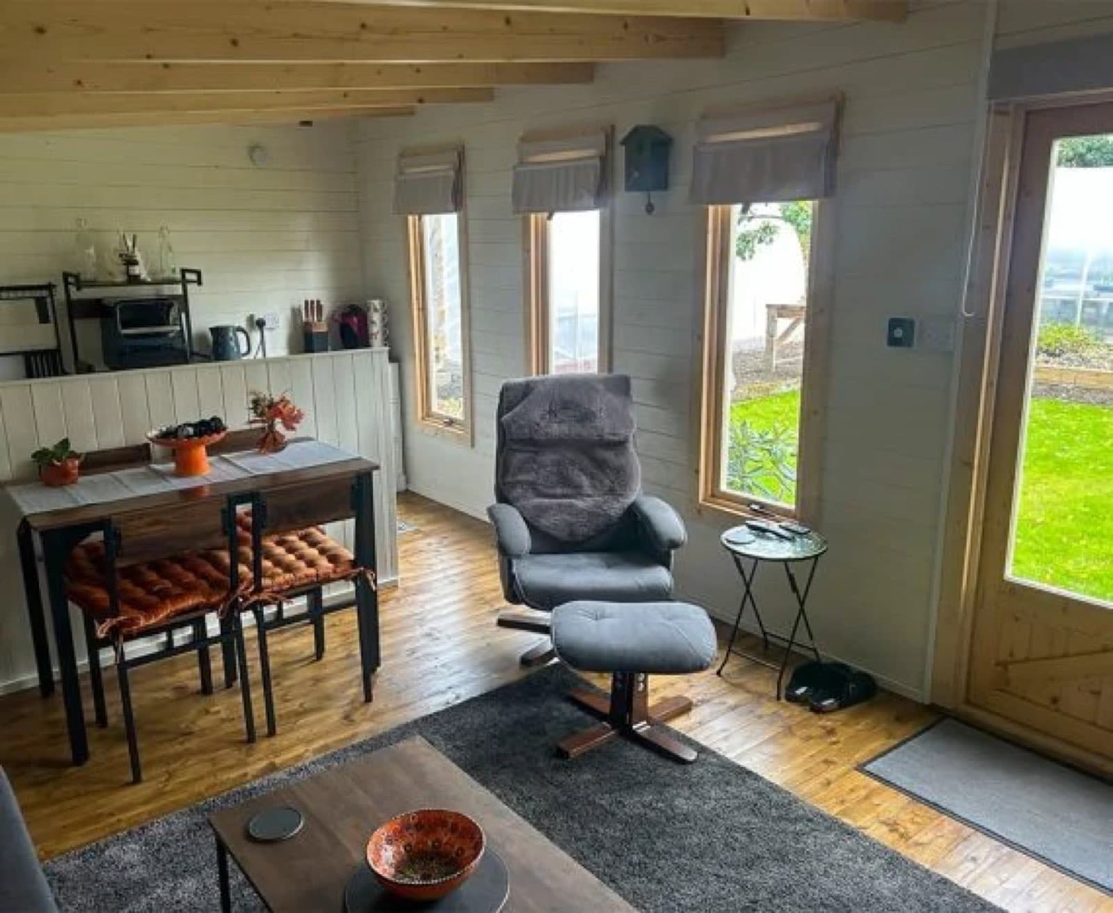 Inside a log cabin living space with a dining table and chairs, a recliner, fitted kitchen units and blinds at the windows