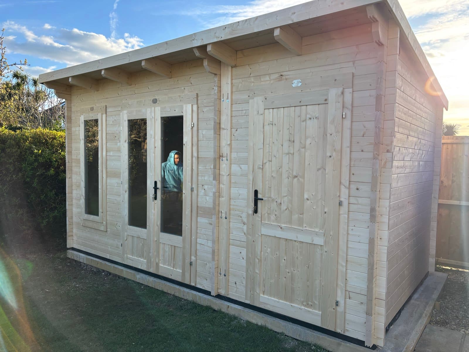 Untreated pent-roof log cabin with glazed double doors, a side window and a separate store door, on a lawn