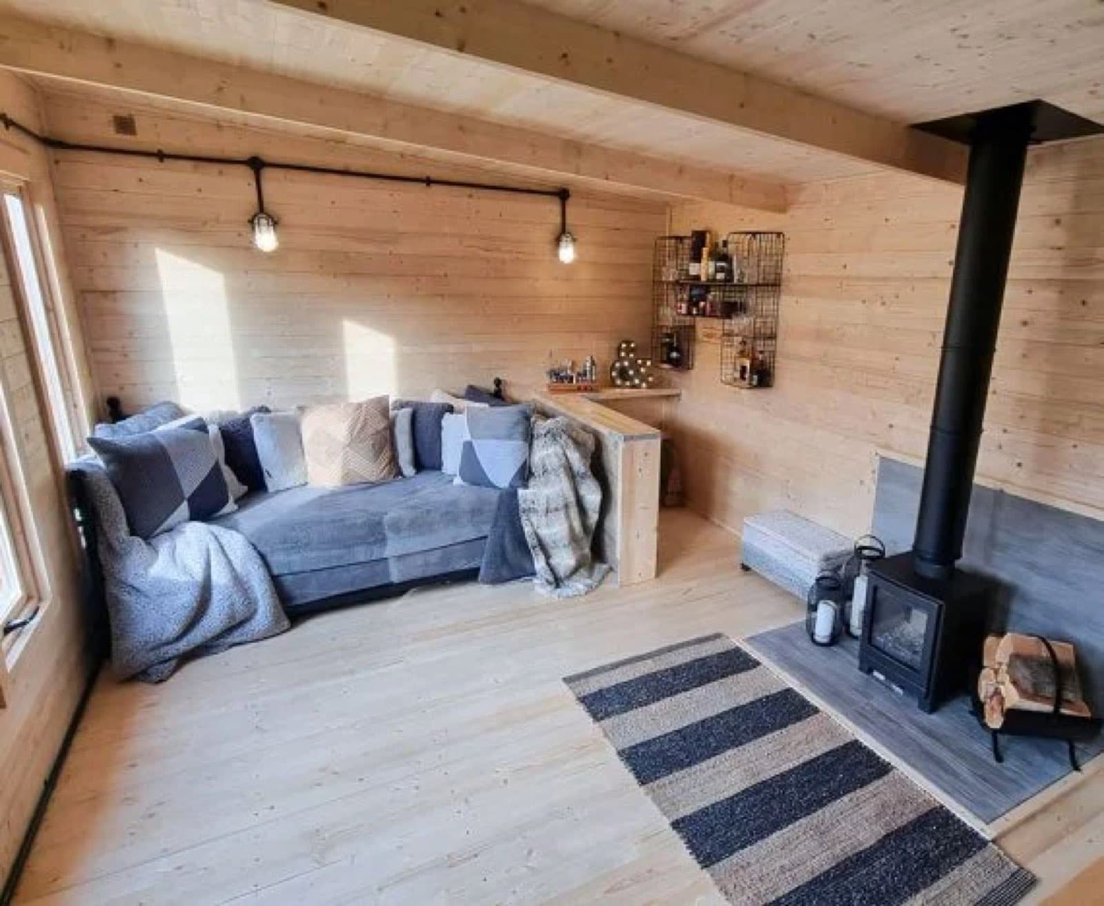Inside a log cabin with a wood burning stove, a grey corner sofa, festoon lights and a striped rug