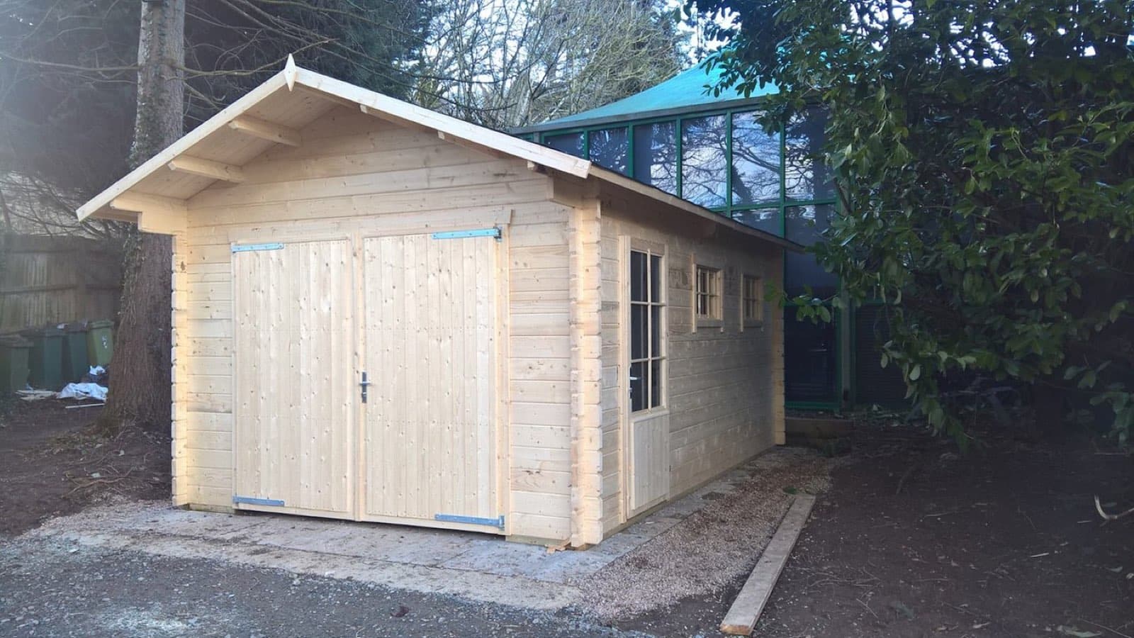 Newly built untreated log garage with an apex roof and double timber doors, next to a greenhouse