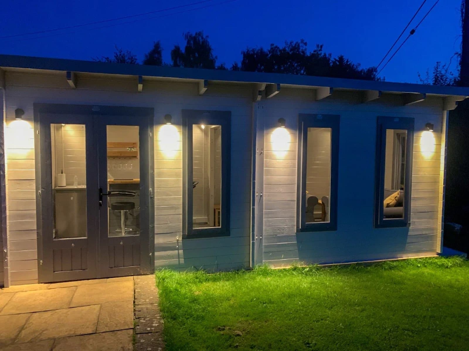 Pale grey multi-room log cabin lit by wall lights at dusk, with a glazed door and three windows along the front