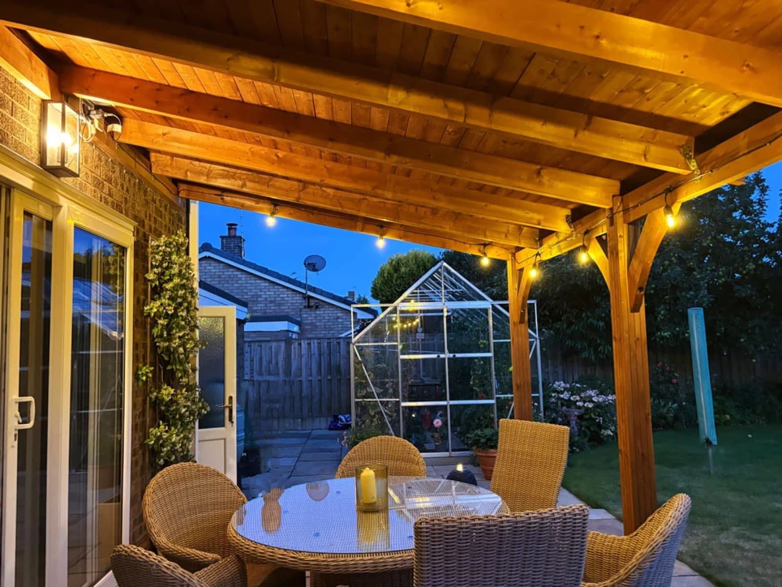 Oak-framed pergola over a patio at dusk, lit with festoon lights, with a rattan dining set underneath and a greenhouse beyond
